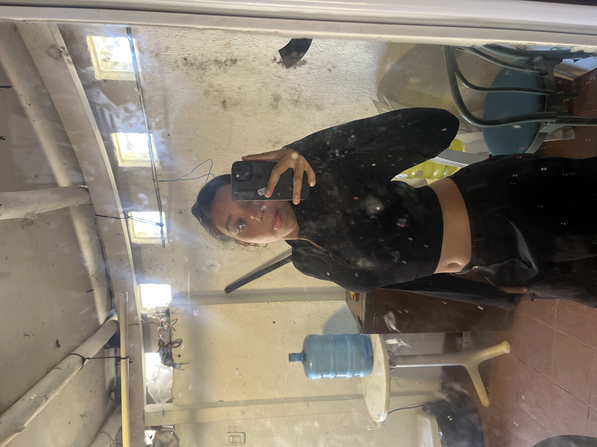 A young woman is captured in a mirror selfie, holding a black smartphone to take the picture. She has dark hair, styled away from her face, and her tongue is slightly out in a playful expression. She is wearing a black long-sleeved crop top that exposes her midriff, paired with dark bottoms.
The setting is an indoor space in Porlamar, Venezuela, reflected in a noticeably dirty and smudged mirror. The room appears to be a functional or utilitarian area, possibly a storage room or workshop, characterized by exposed concrete walls and ceiling beams. Natural light streams in from several small, square openings in the ceiling or upper wall, indicating it is daytime.
In the reflection, a large blue water cooler bottle sits on a light-colored stand or table. Part of a blue chair with metal legs is also visible. The background shows exposed pipes, wires, and general clutter against light-colored walls and a brownish-red tiled floor. The condition of the mirror itself, with numerous specks and streaks, slightly obscures the clarity of the reflection. There is no visible text in the image.