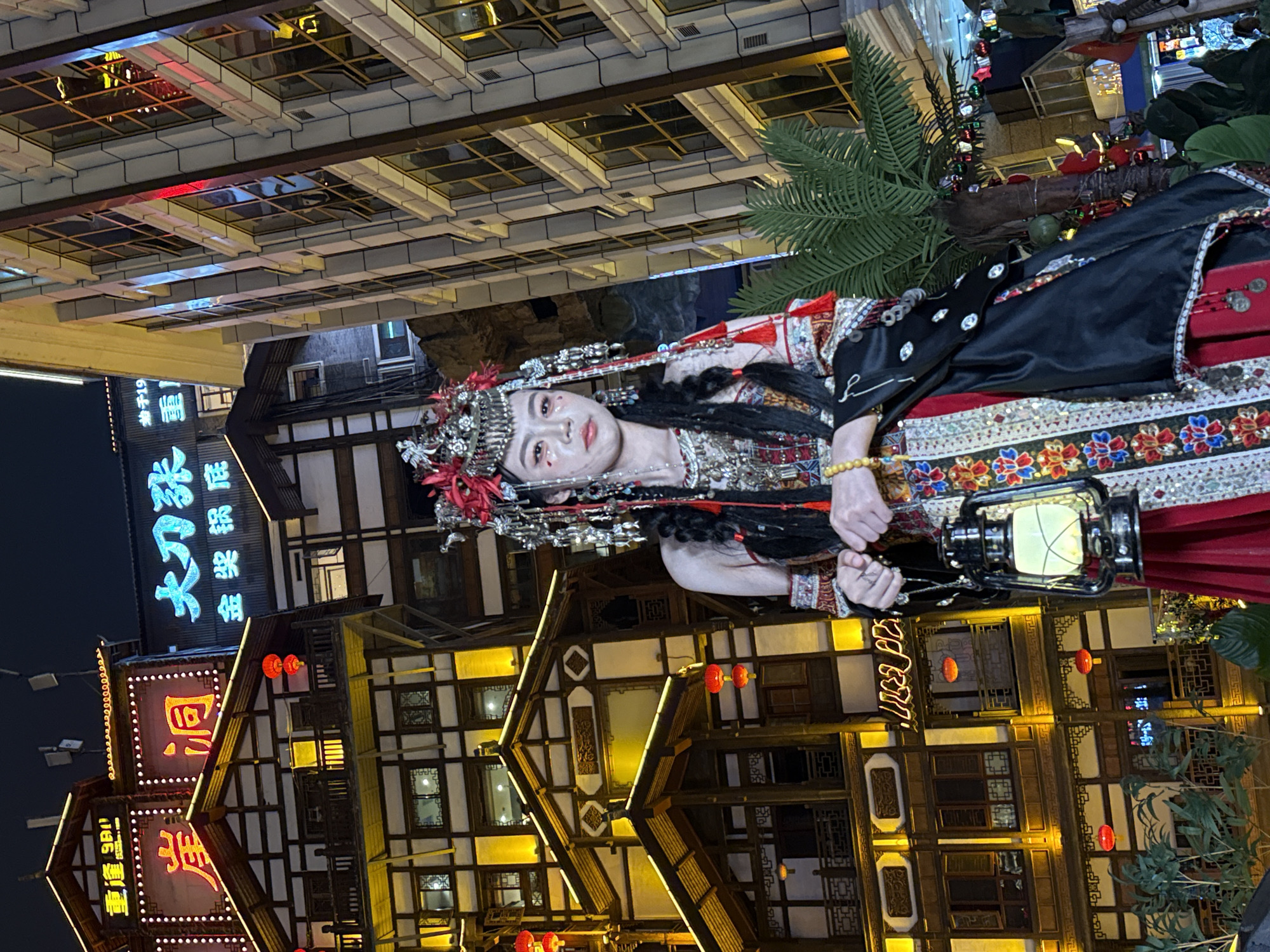 A young woman in elaborate traditional attire is depicted in an evening street scene in Yuzhong District, China. She is the central subject, looking directly at the viewer with a composed expression. Her costume features a black bodice over a richly embroidered garment with red and multi-colored patterns, adorned with metallic accents and tassels. She wears an intricate silver-toned headpiece decorated with red plumes and numerous dangling ornaments. Her makeup includes detailed eye embellishments, and she has long dark hair. On her left wrist, she wears a beaded bracelet, possibly amber. She holds a lit, dark-colored lantern, which casts a warm glow.
Behind her, several multi-story buildings with traditional Chinese architecture are brightly illuminated with warm yellow lights, highlighting their intricate wooden frameworks and numerous windows. The setting appears to be a bustling commercial or cultural area at night. A large palm tree is visible to the right of the woman.
Visible Chinese text on neon signs on the buildings includes "大欢喜火锅" (Dàhuānxǐ Huǒguō, meaning "Great Joy Hotpot") and partial characters like "回" (huí, return) and "廊" (láng, corridor/gallery), suggesting restaurants or shops. The overall scene suggests a cultural display, performance, or a themed street experience.