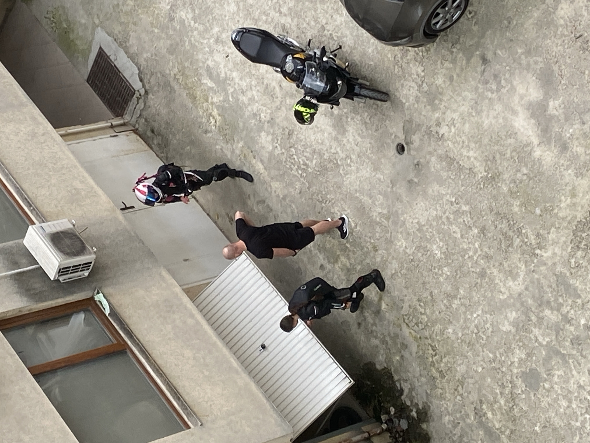 An aerial view depicts an open, concrete courtyard in Varna, Bulgaria, flanked by multi-story buildings. One building has an open window with a visible air conditioning unit. Parked in the courtyard are a black motorcycle, a black helmet, and a portion of a gray car. Three individuals are present: one wearing a motorcycle racing suit and helmet, one casually dressed in black shorts and a black t-shirt, and one in motorcycle attire. Their actions and interactions are unclear from this perspective. A metal grate is visible near one building.