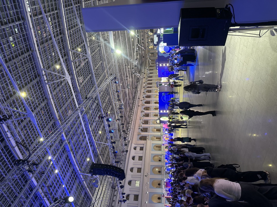The scene depicts a large indoor event taking place at Gostiny Dvor in Moscow, Russia. The venue is characterized by a combination of historical architecture on the left and in the background, featuring light-colored walls, multiple arched windows, and columns, contrasted with a modern event infrastructure overhead.
The high ceiling reveals an exposed metal truss system from which numerous stage lights, spotlights, and a large black line array speaker system are suspended. The overhead lighting casts a predominant blue ambient hue across the space, supplemented by localized white, yellow, and multi-colored lighting from various displays.
Approximately 100-200 individuals, mostly young adults, are present. A dense crowd is congregated or queuing on the far left of the image. Other attendees are scattered throughout the expansive, light-colored, reflective floor, walking, standing, and observing various modern exhibition booths and abstract illuminated displays in the mid-ground. These displays feature vibrant blue, purple, green, and white lighting with contemporary designs, including rectangular panels, curved structures, and circular elements.
In the lower right foreground, a black PA speaker mounted on a stand is visible next to a tall, light blue vertical event pillar. People are generally dressed in casual to smart-casual attire. Their actions indicate general attendance at an event, with no specific organized activities or urgent interactions immediately evident.
