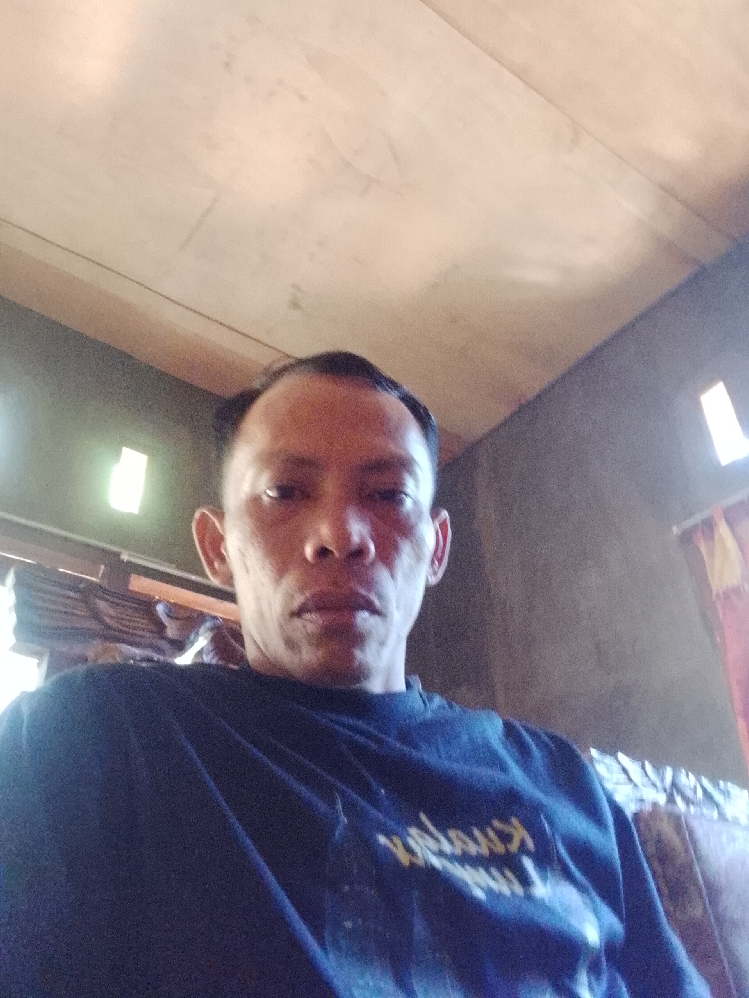 A man with short, dark hair and light skin takes a selfie indoors. He is wearing a dark blue t-shirt with yellow text on the front. The background consists of a wooden ceiling, a grey wall, and two small windows with light emanating from them. There are also some wooden structures visible behind him. The image quality is low, with visible grain and blur. The specific location in Madukoro, Indonesia, is not discernible from the image.