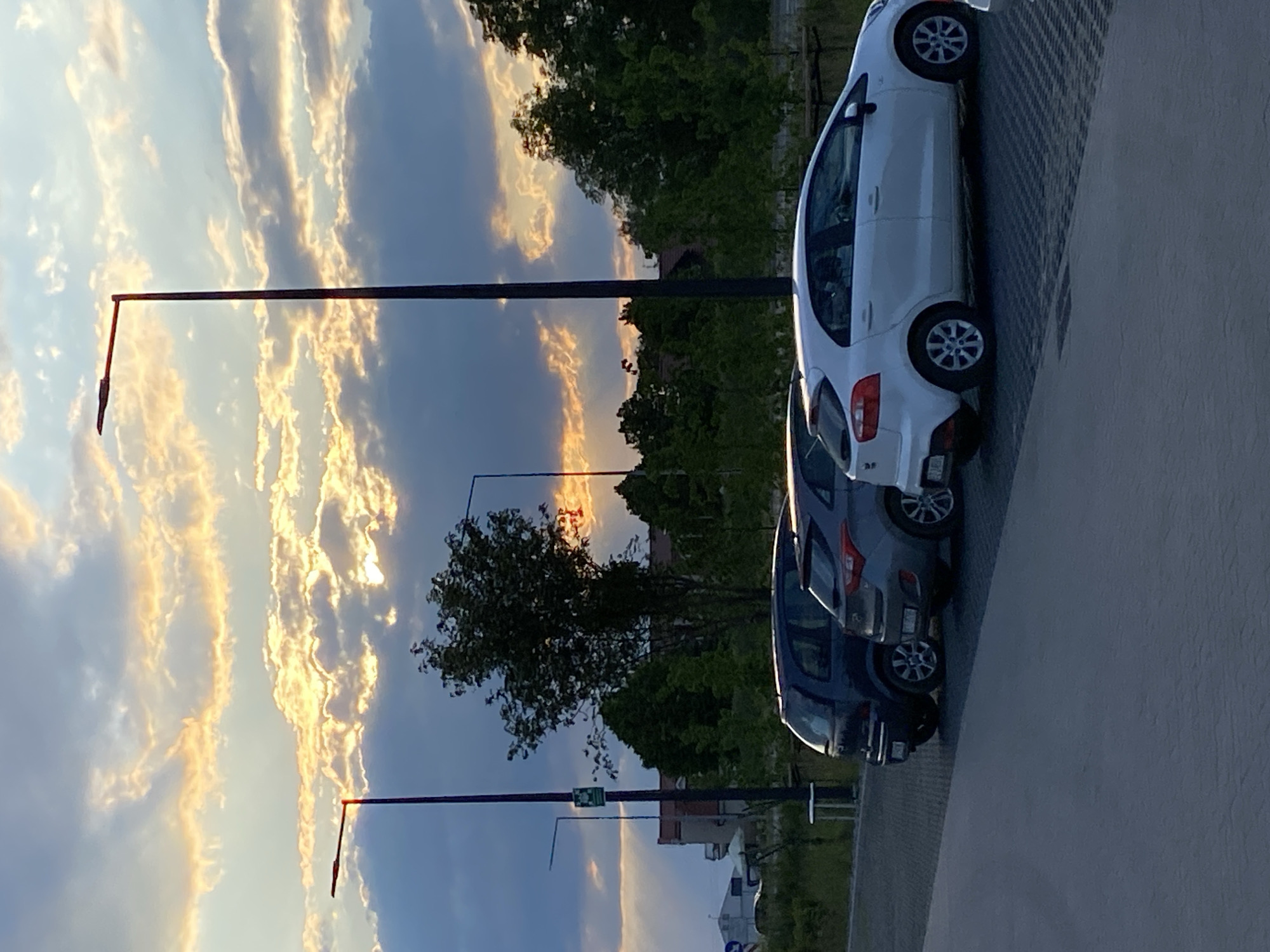 The image shows three parked cars (white, gray, and blue) on a concrete parking area, presumably in the city of Buk, Poland. Background includes trees, sunset, street lights, and partially visible buildings. There are no people depicted in the image.