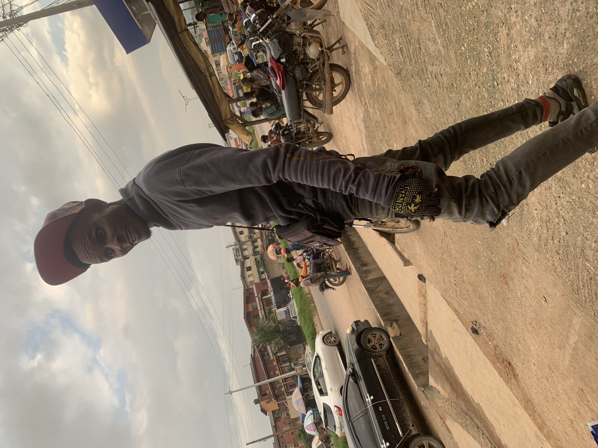 A young Black man wearing a red baseball cap, a grey hoodie, and ripped black jeans stands in what appears to be a busy market area. He is looking to his left, with his mouth open in a surprised expression. His right hand is visible, wearing a black glove with yellow lettering. Behind him, a street scene unfolds with several motorcycles, cars, and buildings. A white sedan is parked on the left, and a black sedan is in the foreground. The sky is overcast with clouds. The man's posture and the surrounding environment suggest a dynamic, possibly chaotic, urban setting in Shagamu, Nigeria.