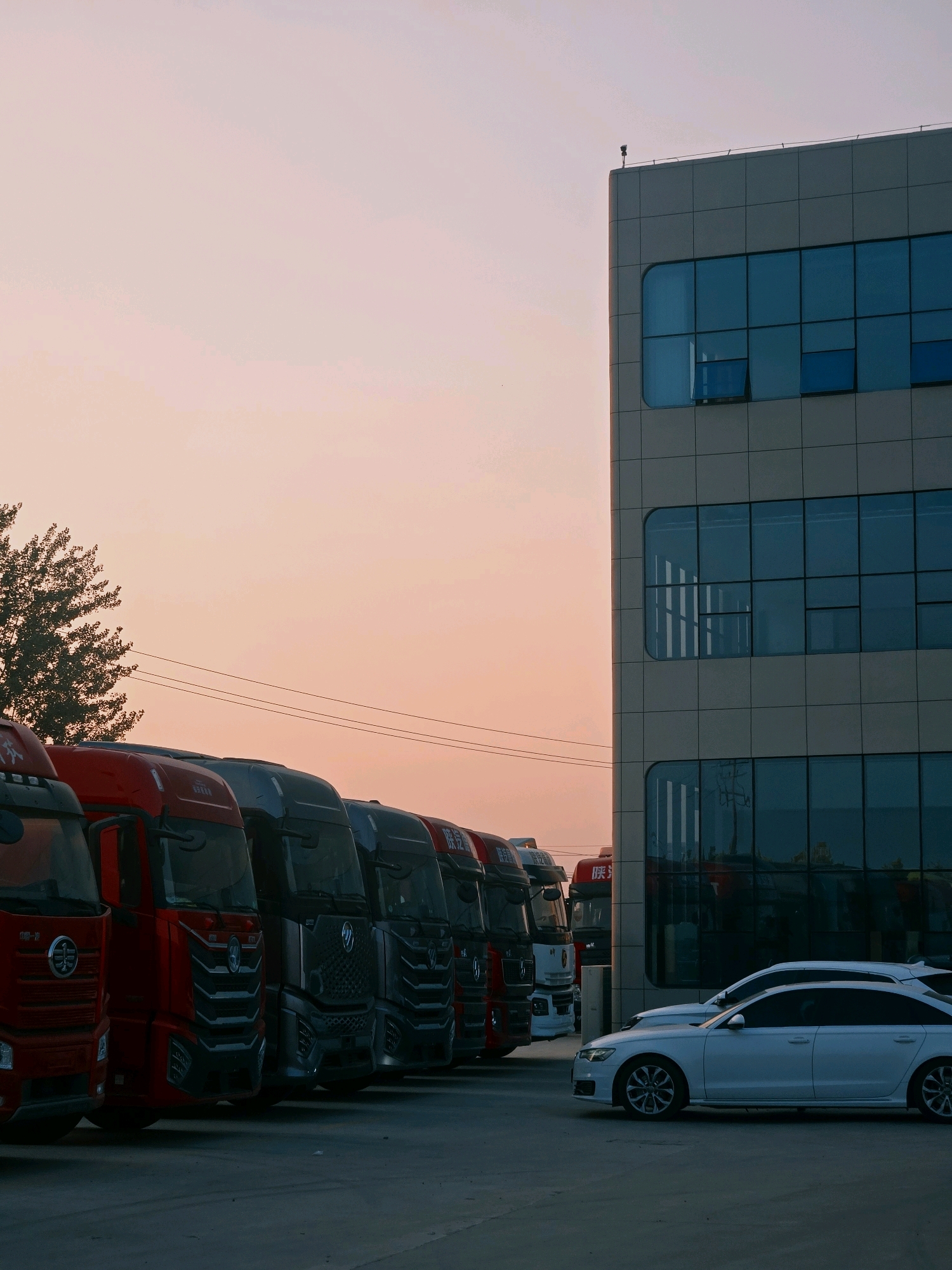 In Liangshan County, China, a fleet of commercial trucks are parked in a lot outside a modern, multi-story building. The trucks, mostly red and gray, are lined up in neat rows, their headlights and grilles illuminated. A white sedan is parked in front of the building, reflecting the building's windows and the soft hues of the twilight sky. The building's facade is characterized by large, rectangular windows that are tinted blue, with some panes open. The sky displays a gradient of pale pink and orange, characteristic of sunset. A tree with dark foliage is visible to the left of the trucks, and power lines stretch across the sky.