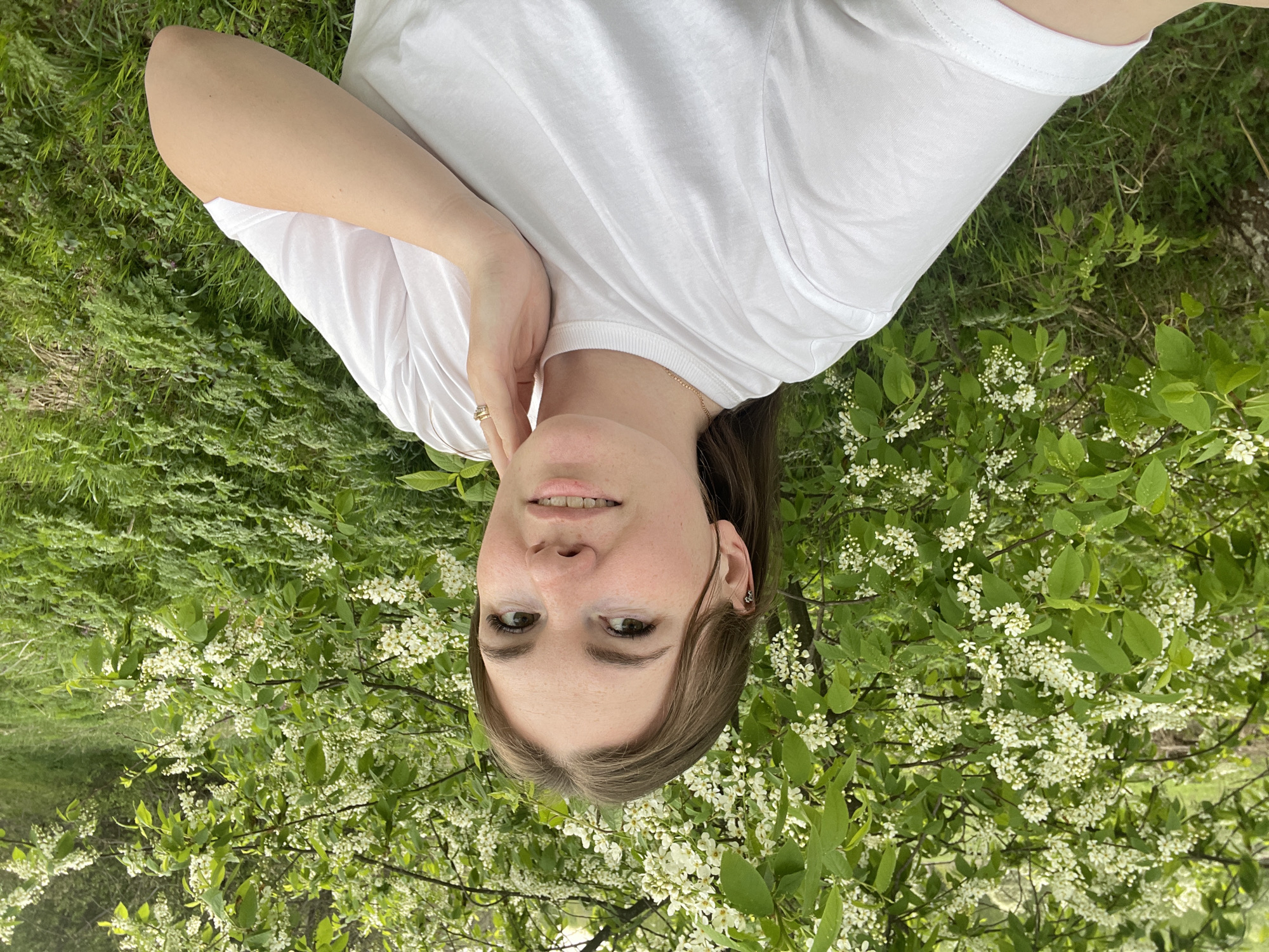 A woman is lying on her back in a grassy area. She is wearing a white t-shirt and has a gold ring on her finger. Her hair is brown and is slightly pulled back. She is surrounded by greenery, including some flowering branches with small white flowers. The exact location in Zhigalki, Russia cannot be determined from the image alone.