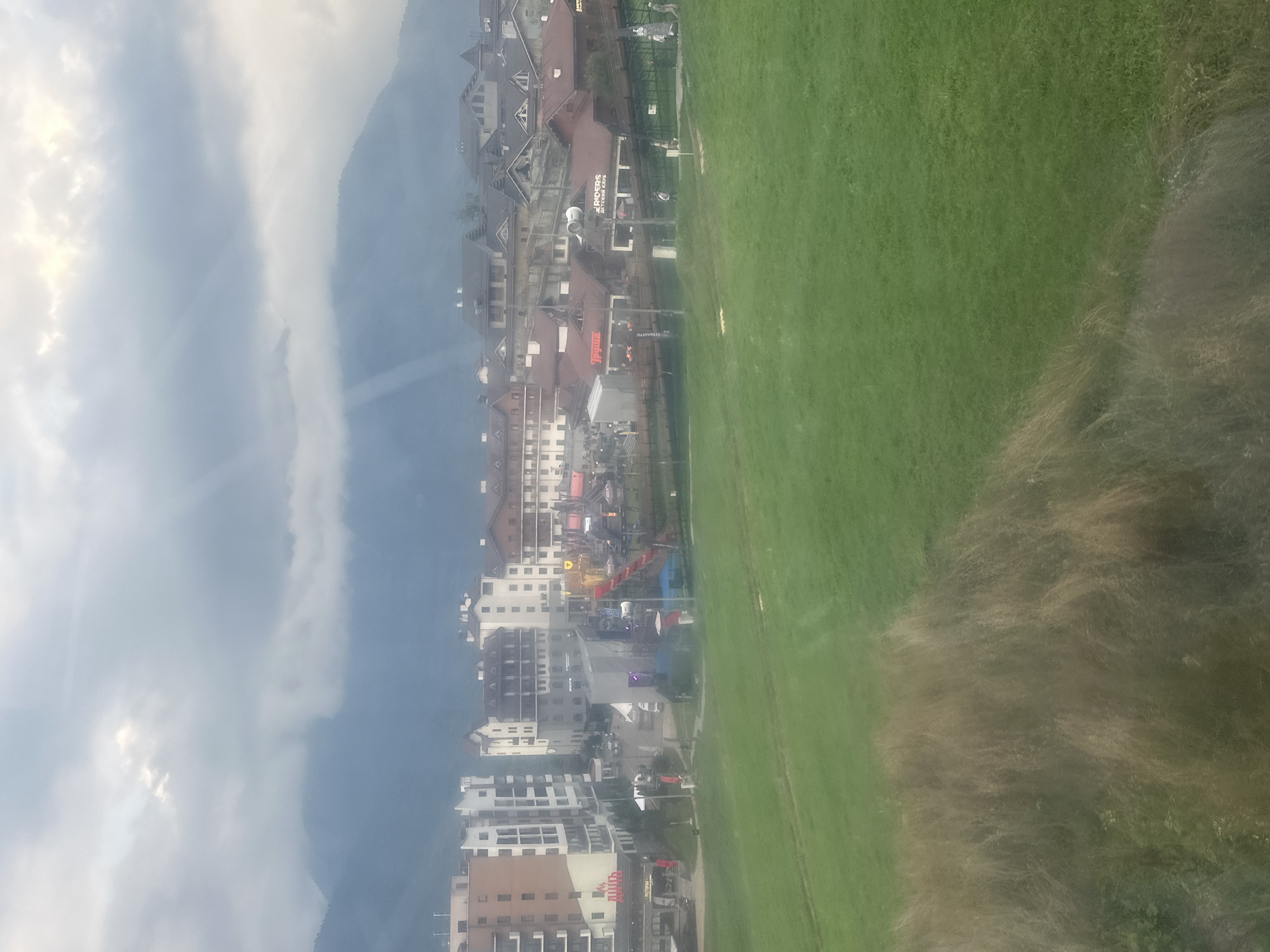 The image depicts a view of the city of Estosadok, Russia, nestled at the base of a mountain range under a cloudy sky. In the foreground, a lush green grassy slope dominates the lower half of the frame, with some dry, brown grasses in the left foreground. Beyond the slope, a collection of multi-story buildings, characteristic of a resort town, are visible. These buildings exhibit a mix of architectural styles, with some featuring modern facades and others older, traditional designs with pitched roofs. Several of these buildings have signage, including one with "Riders" and "Detekativ Cafe" visible. A bustling area with what appears to be an amusement park or event setup is present in the midground, featuring colorful structures and what looks like a stage or performance area. Small groups of people are visible walking around in the midground. The background is characterized by the imposing, mist-shrouded mountains, with rays of sunlight breaking through the clouds, creating a dramatic effect. The overall scene suggests a vibrant resort town in a mountainous region.