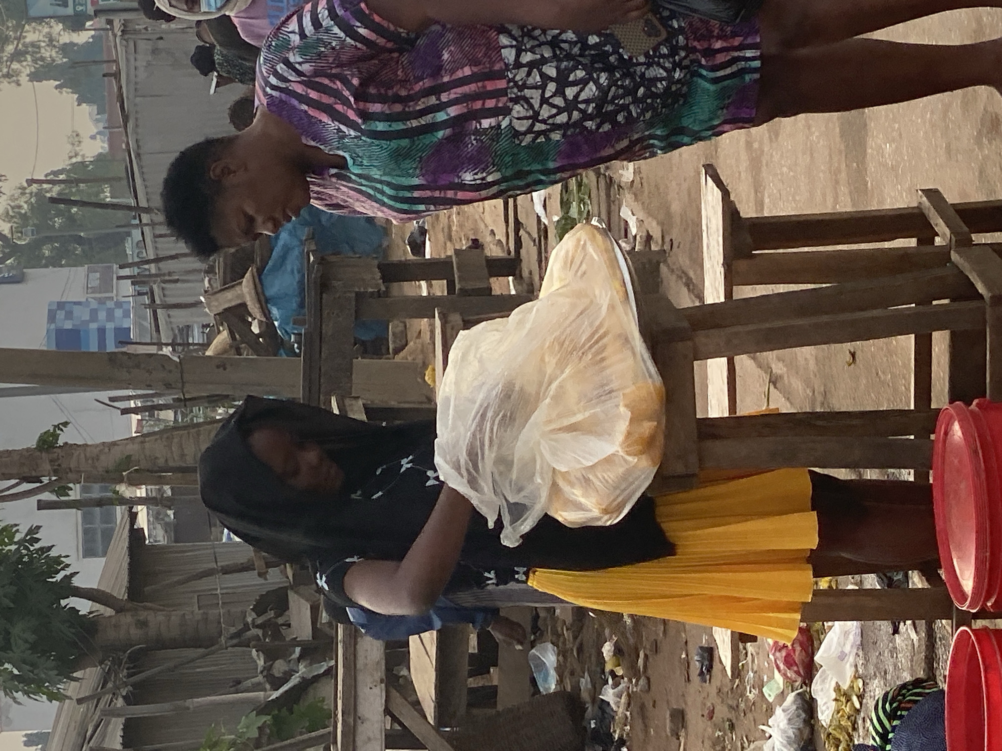 In Sabon Tasha, Nigeria, two women are engaged in a transaction at an outdoor market stall. The woman on the left, wearing a black headscarf and a yellow pleated skirt, is handling a transparent plastic bag filled with what appears to be food items. She is partially obscured by the bag and a wooden stool on which it rests. The woman on the right, dressed in a vibrantly patterned short-sleeved dress, stands facing the first woman, observing the interaction. She is holding a dark object in her right hand, possibly a phone. The environment suggests a bustling, somewhat unkempt market setting with dirt ground, scattered debris, and makeshift wooden stalls and structures in the background. The lighting indicates it is daytime, possibly early morning or late afternoon, with a soft, diffused light.