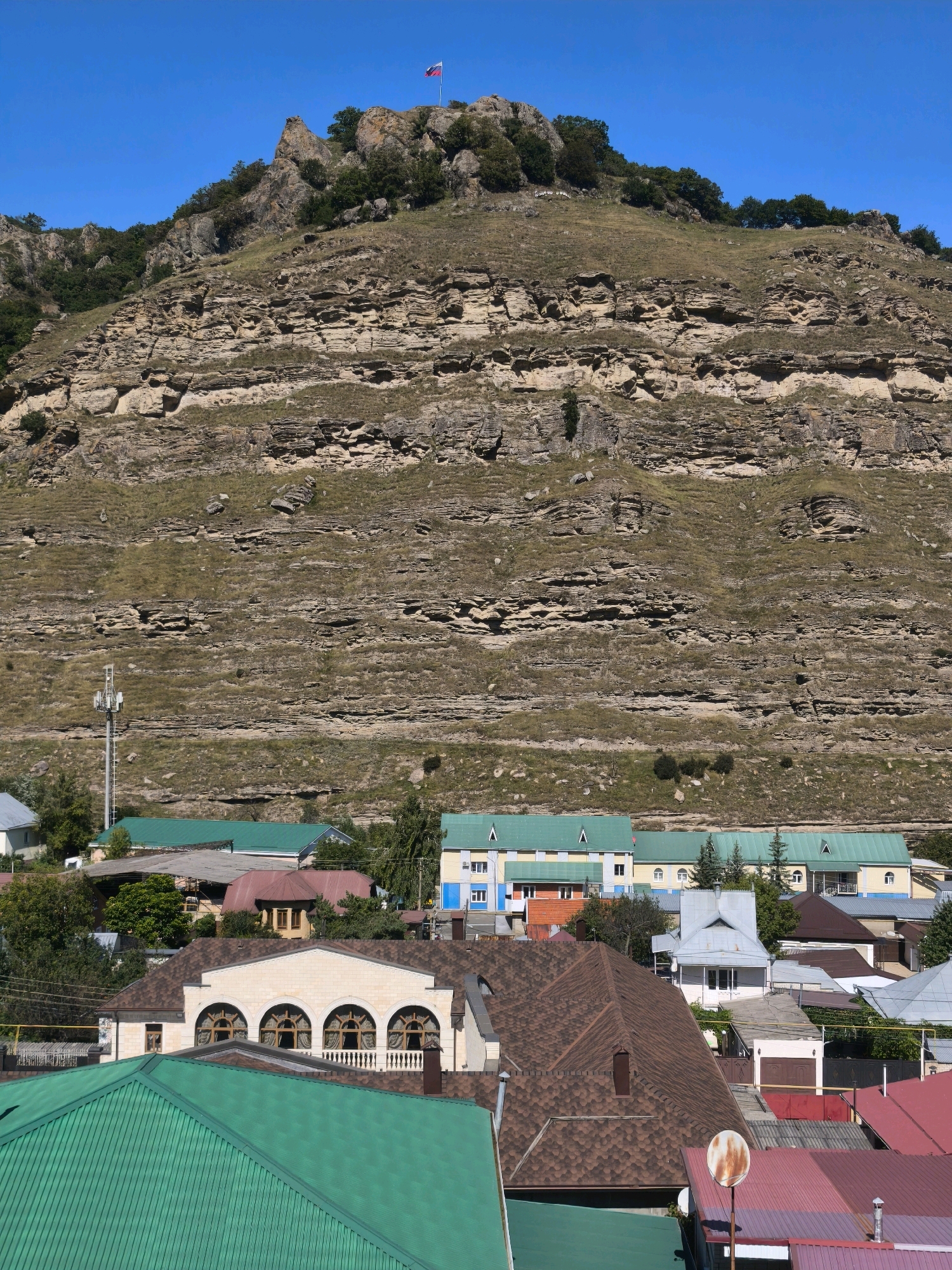 A broad, sunny, and clear blue sky dominates the upper portion of the image, with a slight haze at the horizon. Below the sky, a massive, textured mountain range with prominent horizontal rock strata rises dramatically. The mountain's surface is covered with dry grass and sparse trees, primarily on its upper slopes and crest. A Russian flag flies from a tall pole atop the mountain's peak, serving as a prominent landmark.
In the mid-ground, a cluster of buildings forms a settlement nestled at the base of the mountain. These structures vary in size and design, with several featuring green or turquoise metal roofs. Some buildings have multiple stories, with visible windows and architectural details. A telecommunications tower stands tall among the buildings, its antenna array visible. The buildings appear to be a mix of residential and possibly commercial properties, suggesting a small town or village.
The foreground is filled with the roofs of more buildings, closer to the viewer. These roofs are predominantly made of brown asphalt shingles, with some sections of bright green metal roofing. The architectural styles here are more varied, with some structures featuring arched windows and decorative elements, such as a building with a light-colored facade and multiple arched windows across its upper level. The overall scene depicts a settlement in a mountainous region, likely in Karachayevsk, Russia, given the Russian flag and mountainous terrain characteristic of the Caucasus region.