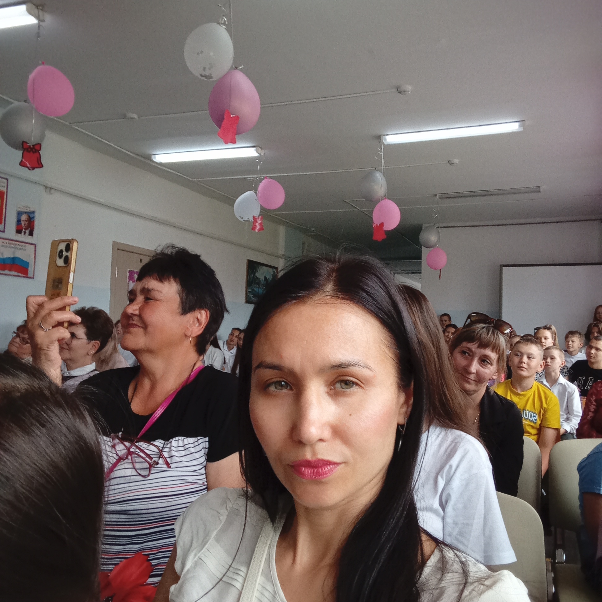 The image shows a medium-angle view of a woman taking a selfie in what appears to be a school auditorium or classroom in Буренка, Russia. The woman has dark hair and is wearing a light-colored top. Behind her, there is a crowd of people sitting in rows of chairs. Some of the individuals are looking at the camera, while others are focused on something else. In the background, there are pink and white balloons hanging from the ceiling. On the wall, there are framed images, including one that appears to be a portrait of Vladimir Putin and the Russian flag. A woman in the background is holding up a gold-colored iPhone, seemingly taking pictures or recording video. The environment is decorated, suggesting a special event or celebration.