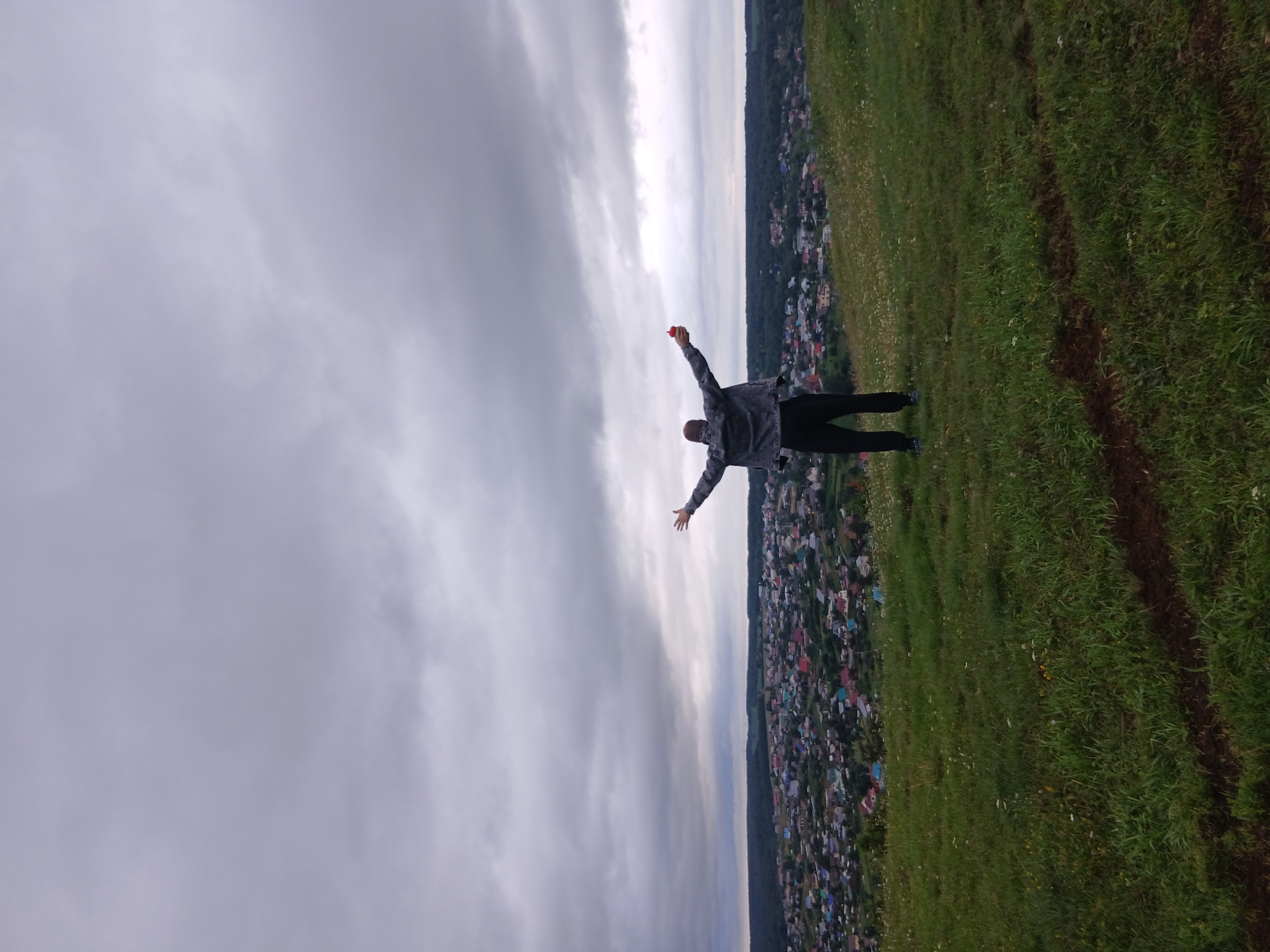 A person stands on a grassy hill, facing away from the camera, with arms outstretched and holding a red object in their right hand. The person is wearing a grey denim jacket and black pants. Below the hill is a sprawling town with numerous houses, surrounded by trees. The sky is overcast with heavy, grey clouds. The location is the city of Olkhovka, Russia.