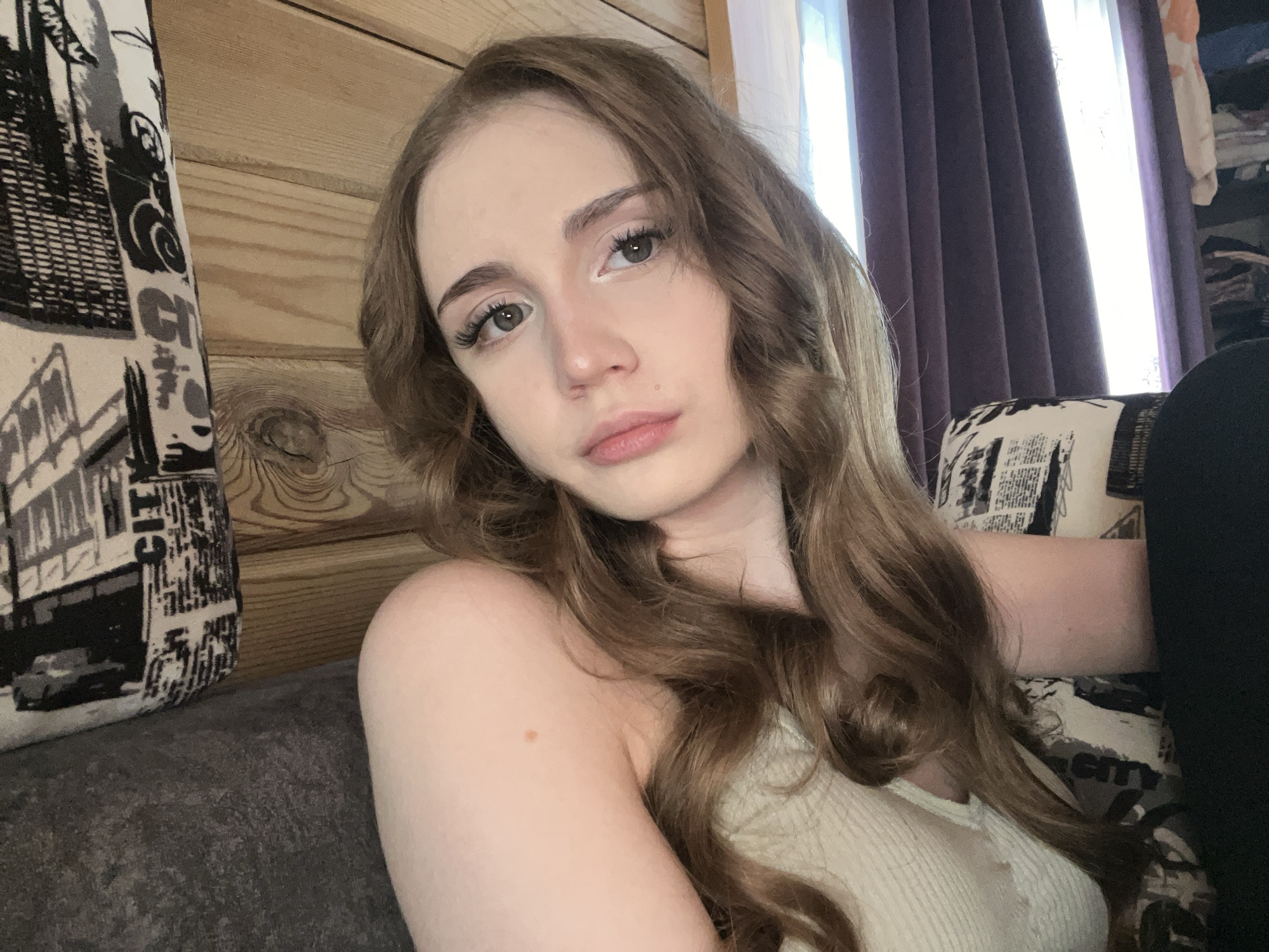 A young woman with long, wavy, light brown hair is the subject of this medium close-up, selfie-style photo. She is looking directly at the camera with a neutral expression, her eyes are green, and she is wearing light makeup. Her skin is fair with a few subtle freckles. She is wearing a light-colored, ribbed tank top.
The background suggests an indoor setting, likely a home. To the left, there is a wooden wall with visible grain. Next to the wall is a couch or chair with a patterned cushion featuring a black and white design that appears to be text and images. To the right, there are dark purple curtains with light filtering through from behind, suggesting a window. Part of a closet or shelving unit with various items is visible behind the curtains on the far right.
The lighting appears to be natural, coming from the window, creating a soft illumination. The time of day is not explicitly indicated, but the light suggests it could be daytime. The overall mood is relaxed and casual. No specific activity or event is taking place beyond the woman posing for the photo.