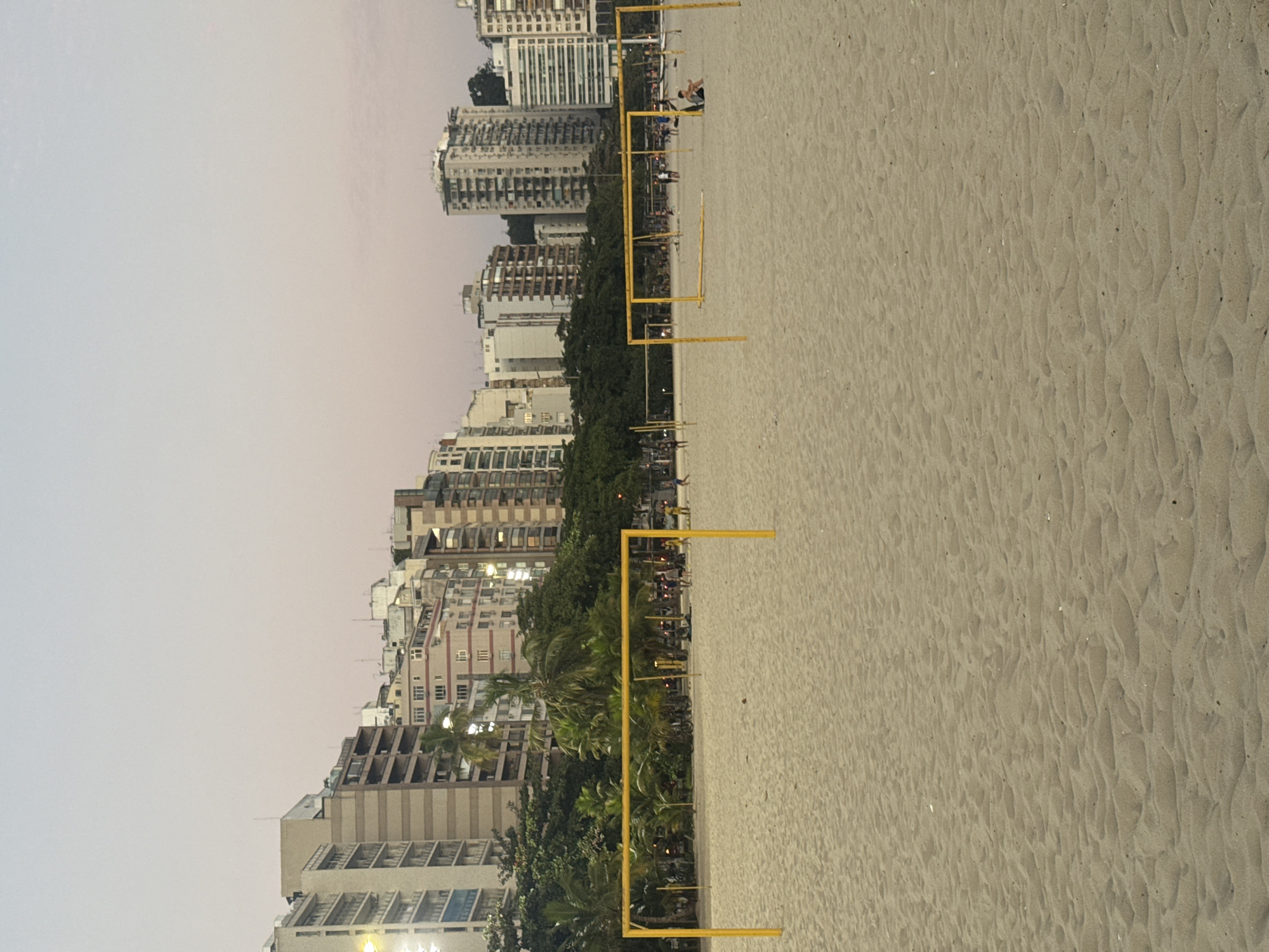 The image depicts a beach scene in Niterói, Brazil, likely taken during the late afternoon or early evening. In the foreground, the sandy beach is marked by several yellow structures that appear to be volleyball nets or goalposts, suggesting recreational activities. Further back, beyond the sandy expanse, is a line of lush green trees and palm trees. Behind the trees, a dense urban landscape of tall buildings rises, characteristic of a city. The sky above is a pale, soft hue, indicating twilight or overcast conditions. A few people are visible on the beach, scattered and engaged in various activities, some near the nets and others in the distance. The overall impression is one of a lively urban beach at the close of day.