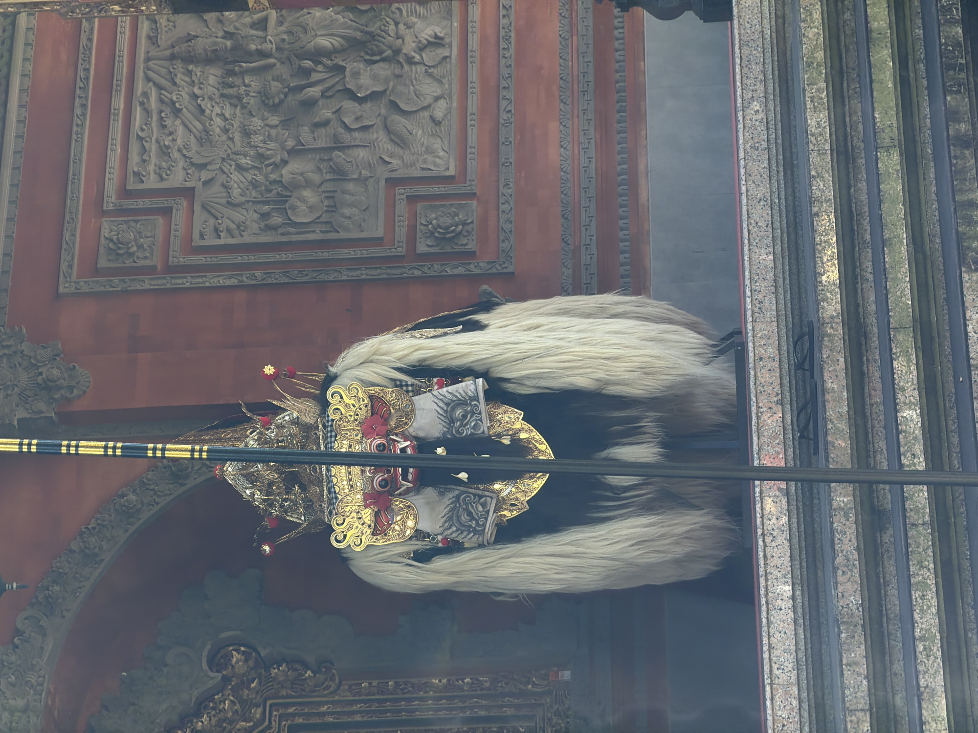 A person wearing a Barong costume is standing on a platform in front of a temple wall. The Barong costume is elaborate, with a golden headdress adorned with red accents and intricate detailing. The costume features a long, shaggy white fur covering its body, with black fur visible underneath. The person's face is concealed by a mask with a red and white design. The background consists of a textured temple wall with carved reliefs, possibly depicting a narrative scene. In the foreground, there are stone steps leading up to the platform. A dark, vertical pole runs through the center of the image, partially obscuring the costume. The overall scene suggests a cultural or ceremonial context within a temple in Baturiti, Indonesia.
