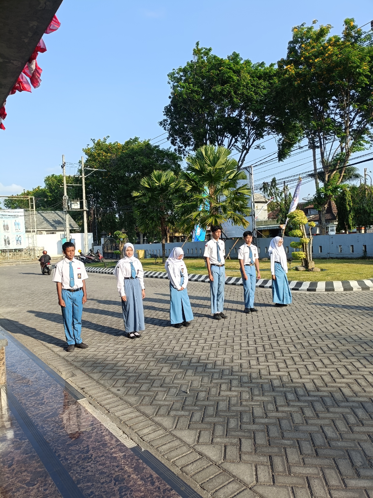 In the city of Kediri, Indonesia, five uniformed students stand in formation on a paved walkway. Two boys, dressed in white shirts, blue ties, and blue trousers, are on the left side of the group. They are followed by three girls, two of whom are wearing white shirts with blue hijabs and blue skirts, while one is wearing a white shirt, blue hijab, and a blue pleated skirt. The students appear to be at attention. The walkway is made of interlocking gray bricks. Behind the students, a grassy area is bordered by a black and white striped curb. Trees with green foliage and some yellow flowers are visible in the background, along with buildings and power lines. A motorcycle can be seen in the distance on the left side of the image. Red and white flags are draped from an overhang in the upper left corner of the frame. The sun casts shadows on the ground, indicating it is likely morning or afternoon.