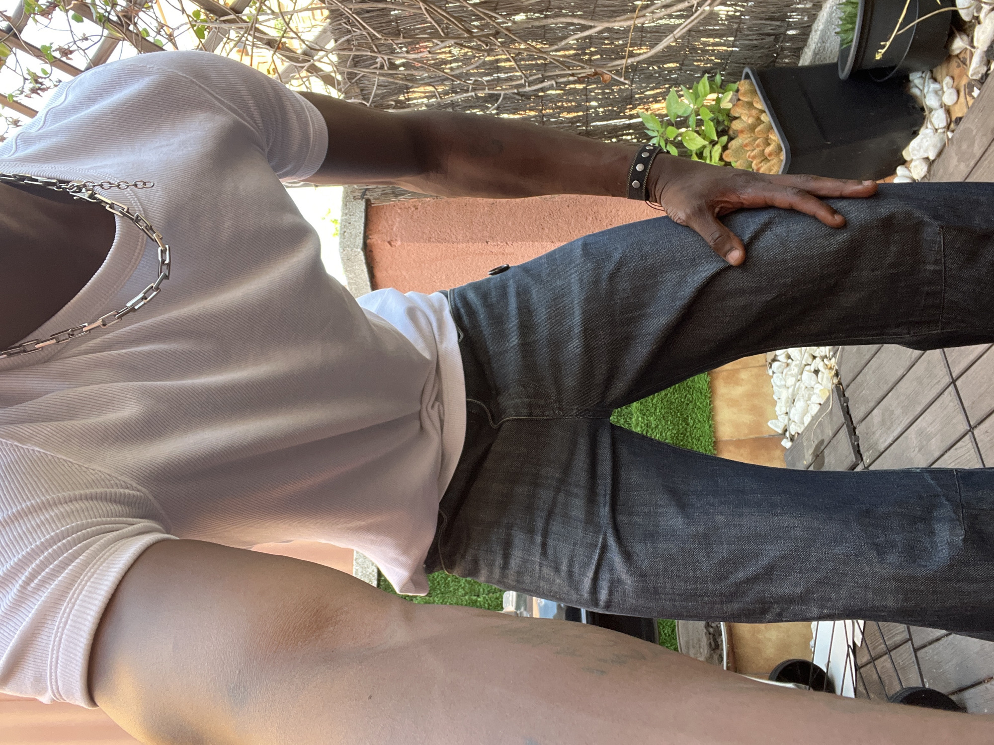 A medium shot of a person from the chest down, positioned outdoors on a wooden surface. The person is wearing a light-colored, short-sleeved ribbed shirt and dark wash jeans. A silver chain necklace is visible around their neck. The person's right hand rests on their right thigh, with a dark studded bracelet on their wrist. In the background, a terracotta-colored wall is partially visible, along with greenery in black planters and a partially visible fence. The location is most likely an outdoor patio or terrace. There is no evidence that this location is in Barcelona, Spain.