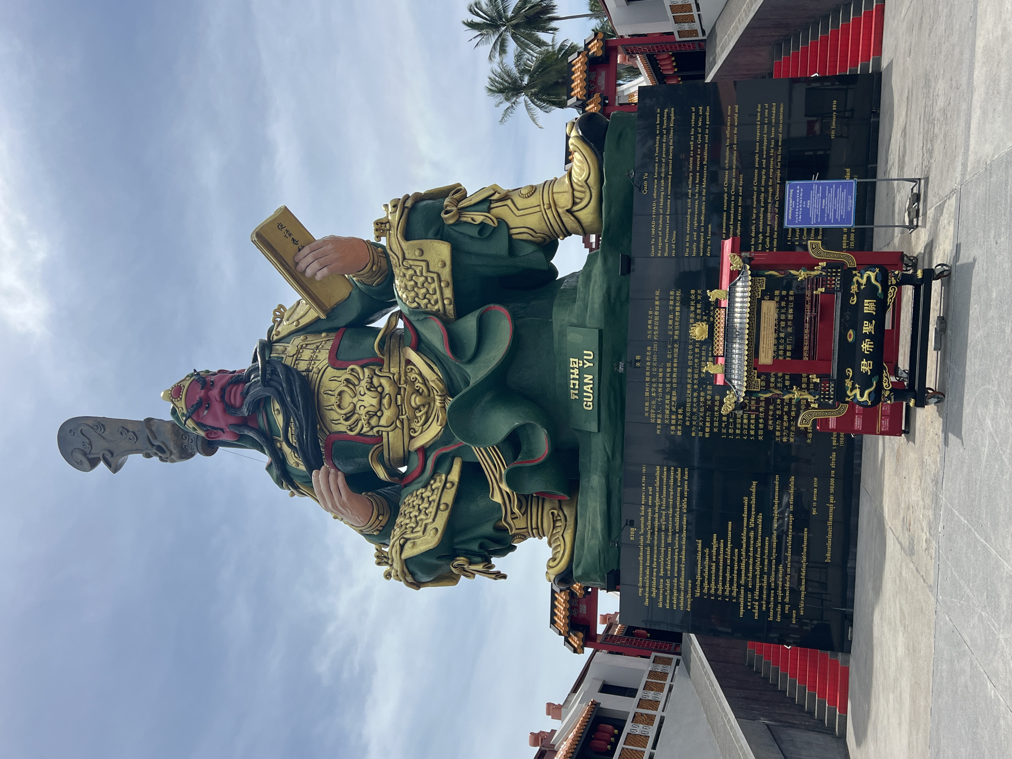 The image shows a large, imposing statue of Guan Yu, a historical figure and military general from the Three Kingdoms period of China. The statue is predominantly green with gold accents, depicting Guan Yu in a seated position, holding a book. He has a red face, a long black beard, and wears elaborate armor and a headdress. The statue is situated in Baan Hua Thanon, Thailand, and is mounted on a large black pedestal with inscriptions in both Thai and English. The English text on the pedestal details Guan Yu's life, his virtues, and his reverence as a God of War. In front of the pedestal, there is a smaller, ornate red and gold incense burner. The background features a bright blue sky with scattered clouds, and on either side of the statue, there are traditional red and gold temple structures. Red stairs lead up to the platform where the statue is located. The overall scene conveys a sense of historical significance and religious veneration.