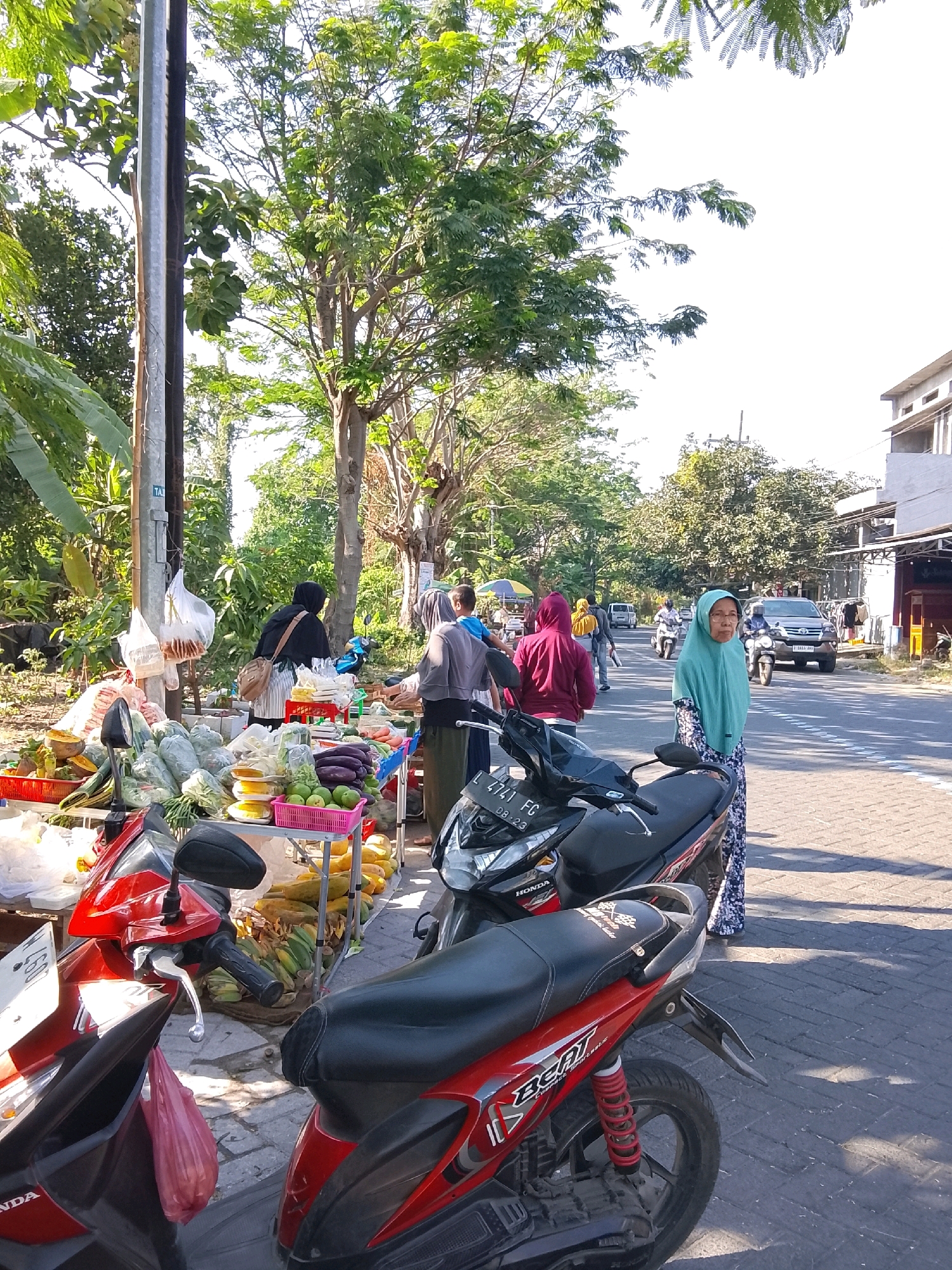 An outdoor, daytime scene depicts a roadside market in Damarsi, Indonesia, with a variety of fresh produce displayed on tables and on the ground. Two red and black motorcycles are parked in the foreground. Several individuals, including women wearing hijabs, are visible at the market, some interacting with the produce and others standing. In the background, a street with vehicles, trees, and buildings suggests a moderately populated urban or suburban area. The lighting indicates it is a sunny day.