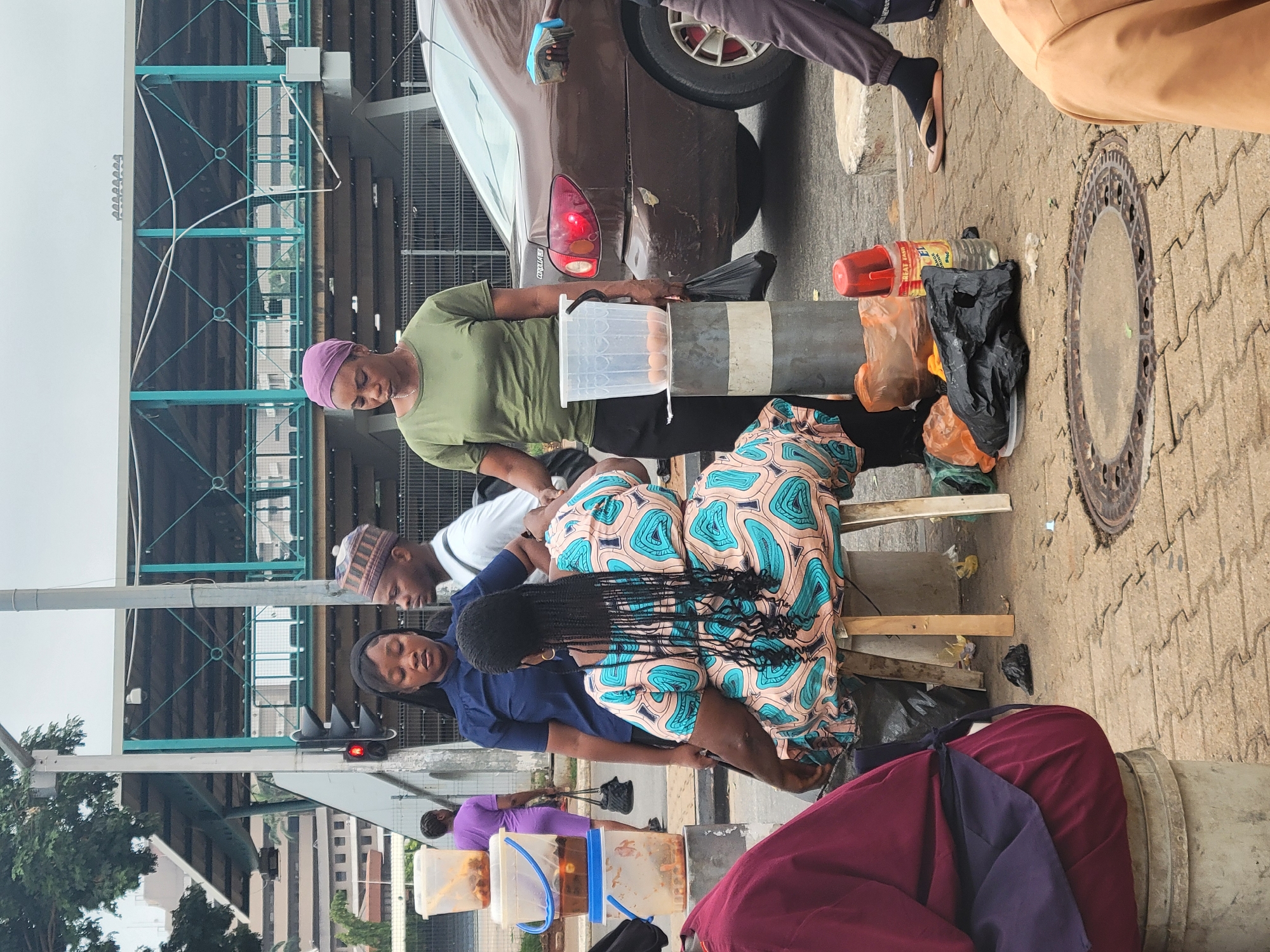 A street scene in Abuja, Nigeria. Three women are sitting on small wooden stools next to a curb. The woman in the middle wears a colorful turquoise and tan patterned dress. One woman is wearing a purple headscarf and a green top and is standing next to a barrier that appears to have eggs on it. To the left is a traffic light and a pedestrian bridge. A dark-colored car is parked beside the scene, with a person reaching into it. Several plastic containers and bags are also present on the ground.