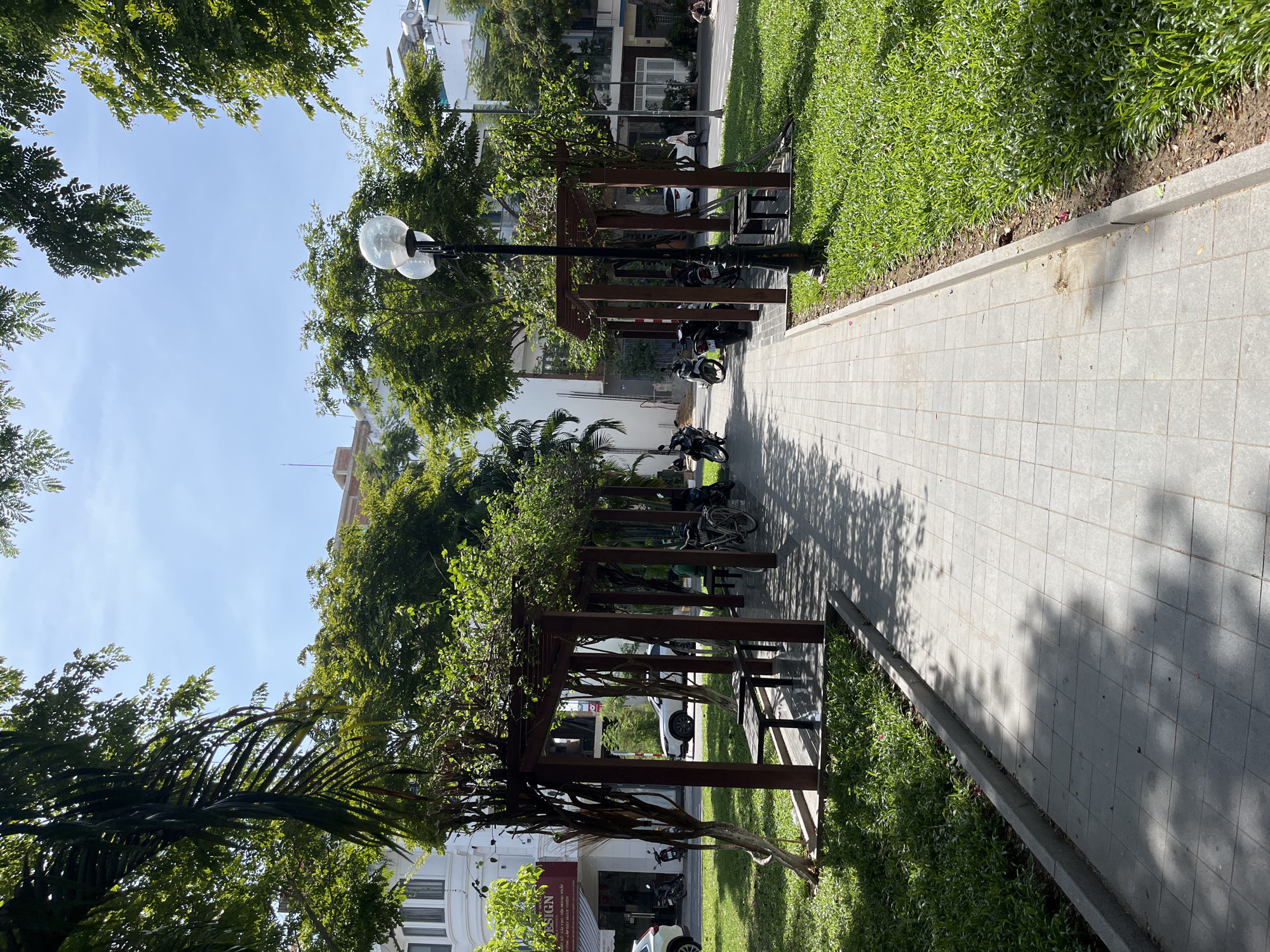 The image depicts a public park area in Nam Nha Trang Ward, Vietnam, featuring a paved walkway with a manicured lawn on the right and a series of shaded pergolas with benches on the left. The walkway is constructed of light-colored square tiles and is bordered by a raised edge and the grassy area.
The pergolas, made of dark wood, create covered seating areas. Benches are visible within some of these structures. Several motorcycles are parked along the side of the walkway, near the buildings that line the street beyond the park. In the background, there are multi-story buildings, some appearing to be commercial establishments with signage.
The environment is lush and green, with mature trees providing shade. Sunlight filters through the leaves, casting dappled shadows on the walkway and the grass. The sky is clear and blue. A street lamp with two globe-shaped lights stands prominently near the center of the frame, casting a shadow. There are a few people visible in the distance, one sitting on the grass and another walking.