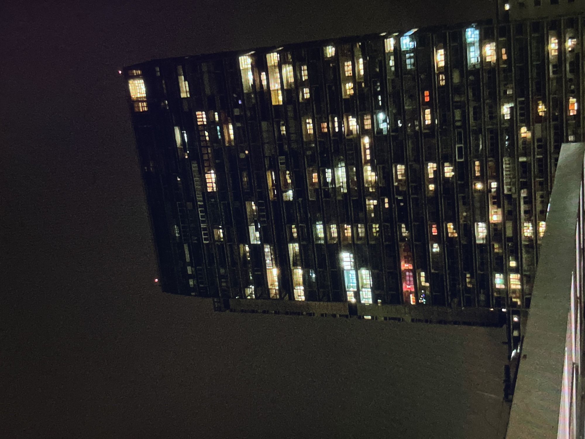 A nighttime view features a tall building with numerous lit windows, suggesting residential or office occupancy. The building's architectural style appears modern with a grid-like façade. The environment is dark, likely urban, with the building's lights contrasting against the night sky. A portion of what seems like a balcony or higher-level structure is visible on the right, offering a partial vantage point of the building.