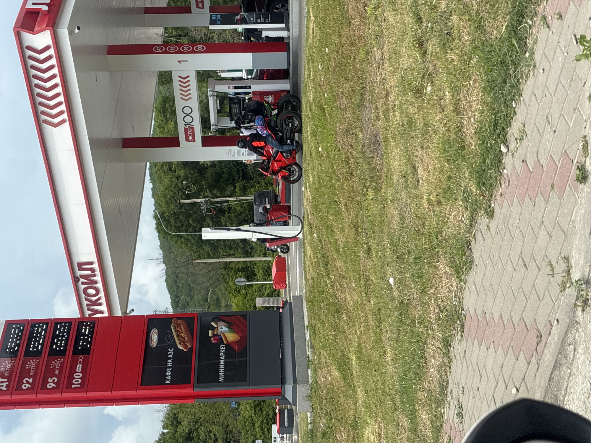 In Arkhipo-Osipovka, Russia, a gas station with red and white branding is visible. A prominent red sign displays fuel prices, with digits "92," "95," and "100" next to the word "ЭКТО," indicating different fuel types. Below the prices, images of a hot dog and coffee, and a minimarket are advertised. The gas station canopy features a white roof with red chevron-like designs and the word "УКОЙЛ" in large letters. Two motorcycles are parked at a gas pump. One is a red sportbike with a rider wearing a black helmet, orange jacket, and blue jeans. The other, a green motorcycle, is parked behind the red one, with another rider wearing a helmet. The scene is set against a backdrop of green trees and a cloudy sky. The foreground shows a paved walkway and a grassy area.