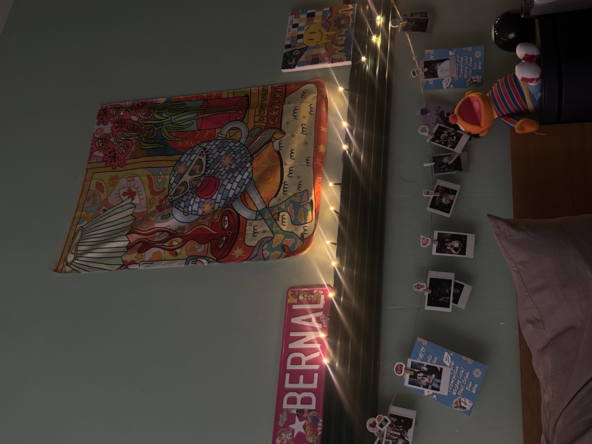 The image shows a bedroom wall decorated with various items. On the left, there's a tapestry with colorful abstract designs. Above it is a small painting with a smiley face. A string of fairy lights runs vertically along the wall, next to a pink sign that says "BERNAL". Mini clothespins hold up a string of small photographs and decorative cards, including one that says "PARTY" and lists various cocktails and drinks. To the right, part of a bed with a lavender pillow is visible, along with a small toy and a black coffee maker on a wooden headboard. The location in Moscow, Russia is not discernible from the image.