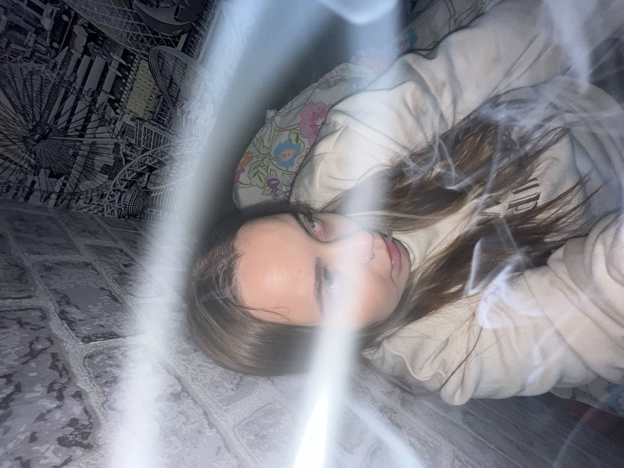 The image captures a young woman lying down, looking directly at the camera in an indoor setting in Novopokrovskaya, Russia. Her head is tilted to the left, and she has long, light brown hair, fair skin, and a nose piercing on her right nostril. Her eyes appear to have a reddish tint, possibly a red-eye effect from a flash. She wears a light-colored, possibly beige or off-white, hooded sweatshirt, with some dark, partially visible text on its front, though it's obscured by lighting effects.
The surrounding environment features a dark-toned wallpaper or mural to her left, displaying intricate, monochromatic designs of what appears to be a cityscape or amusement park with structures resembling Ferris wheels and buildings. This wallpaper has a slightly textured or glittery finish. Underneath her head, a pillow with a colorful floral pattern (pink, blue, and green flowers on a light background) is visible. To the bottom left of the frame, a gray surface with a brick-like pattern serves as part of the immediate foreground or floor.
The photograph is significantly characterized by prominent, bright white light streaks or lens flares that cut diagonally across the image, casting a glow over parts of the subject and background. The scene is illuminated by what appears to be artificial light, with no discernible outdoor light cues. The woman's expression is neutral.