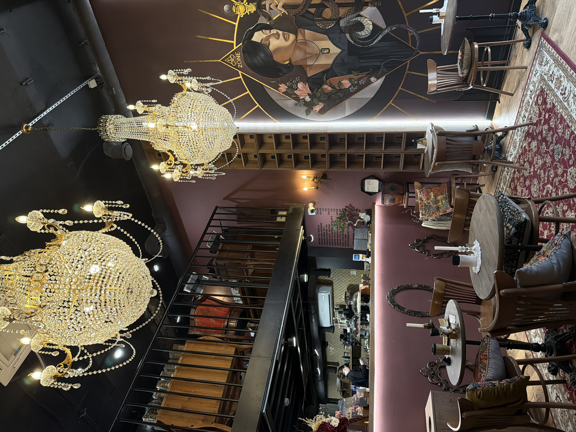 The image shows the interior of a cafe or restaurant, possibly located in Moscow, Russia, given the Cyrillic text on a menu board. The scene is observed from an elevated perspective, with two ornate chandeliers dominating the foreground. The lighting appears dim, enhancing the ambiance.
On the ceiling, there's a large mural depicting a woman surrounded by snakes and zodiac symbols. The interior design features wooden furniture, including tables and chairs, and a staircase with metal railings. A rug with an intricate pattern lies on the floor.
A person can be seen behind the counter, possibly a barista or cashier. The environment appears upscale and gothic-inspired.