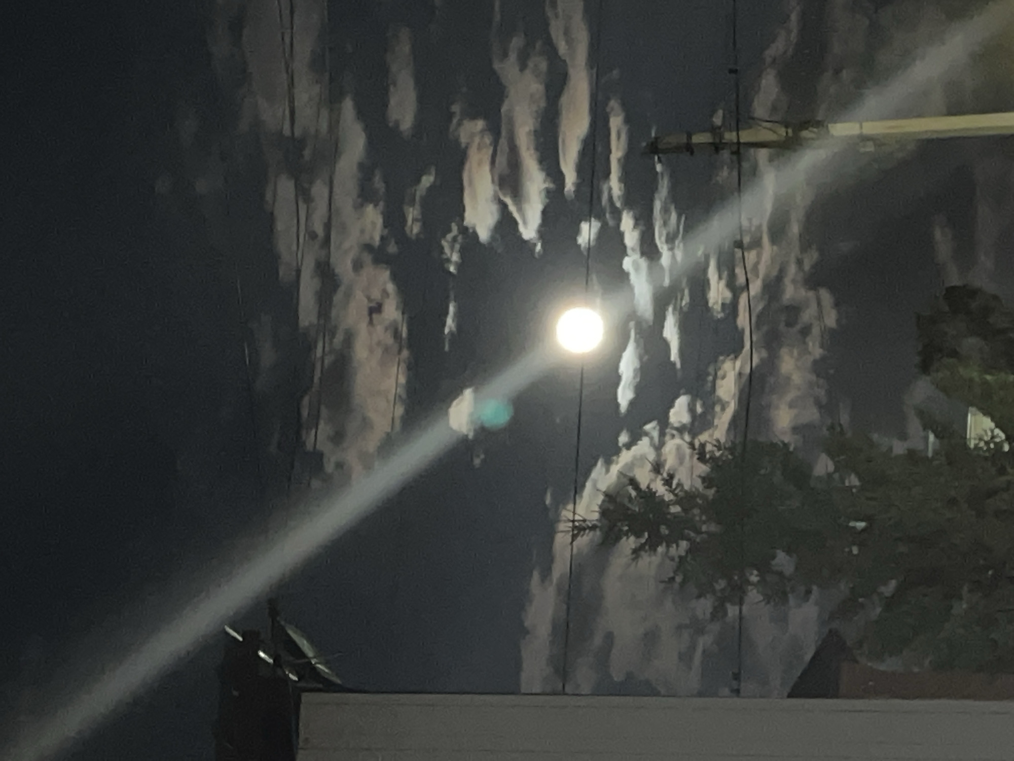 This is a nighttime photograph taken in Saratov, Russia, featuring a full moon partially obscured by clouds. A bright beam of light, possibly from a streetlamp or spotlight, cuts diagonally across the frame from the upper left towards the lower right, illuminating some of the clouds. A solitary evergreen tree is visible in the lower left quadrant of the image. In the background, power lines stretch horizontally across the sky. A utility pole stands on the right side of the image, with its structure illuminated by the overhead light. The scene appears to be in a residential or semi-urban area, with the edge of a building visible in the far left.