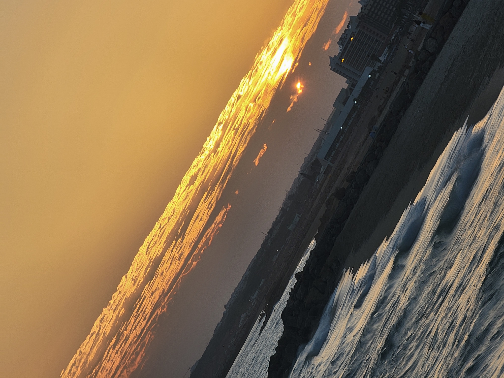 The image depicts a tilted view of a beach scene in Quarteira, Portugal, during what appears to be sunset. The sky is dominated by warm hues of orange and yellow, with a strip of illuminated clouds stretching diagonally across the frame from the lower right to the upper left. The sun, a bright orb, is partially obscured by these clouds, casting a golden light.
In the foreground, the dark blue ocean water is in motion, with white-capped waves reaching towards a rocky shoreline. Beyond the rocks is a sandy beach, populated by several individuals and small groups. Their figures are small and indistinct due to the distance and lighting conditions. Further inland, a series of buildings, including what appear to be hotels, are visible. Some of these structures are tall and modern, with windows reflecting the fading light. The overall impression is one of a coastal city at the end of the day.