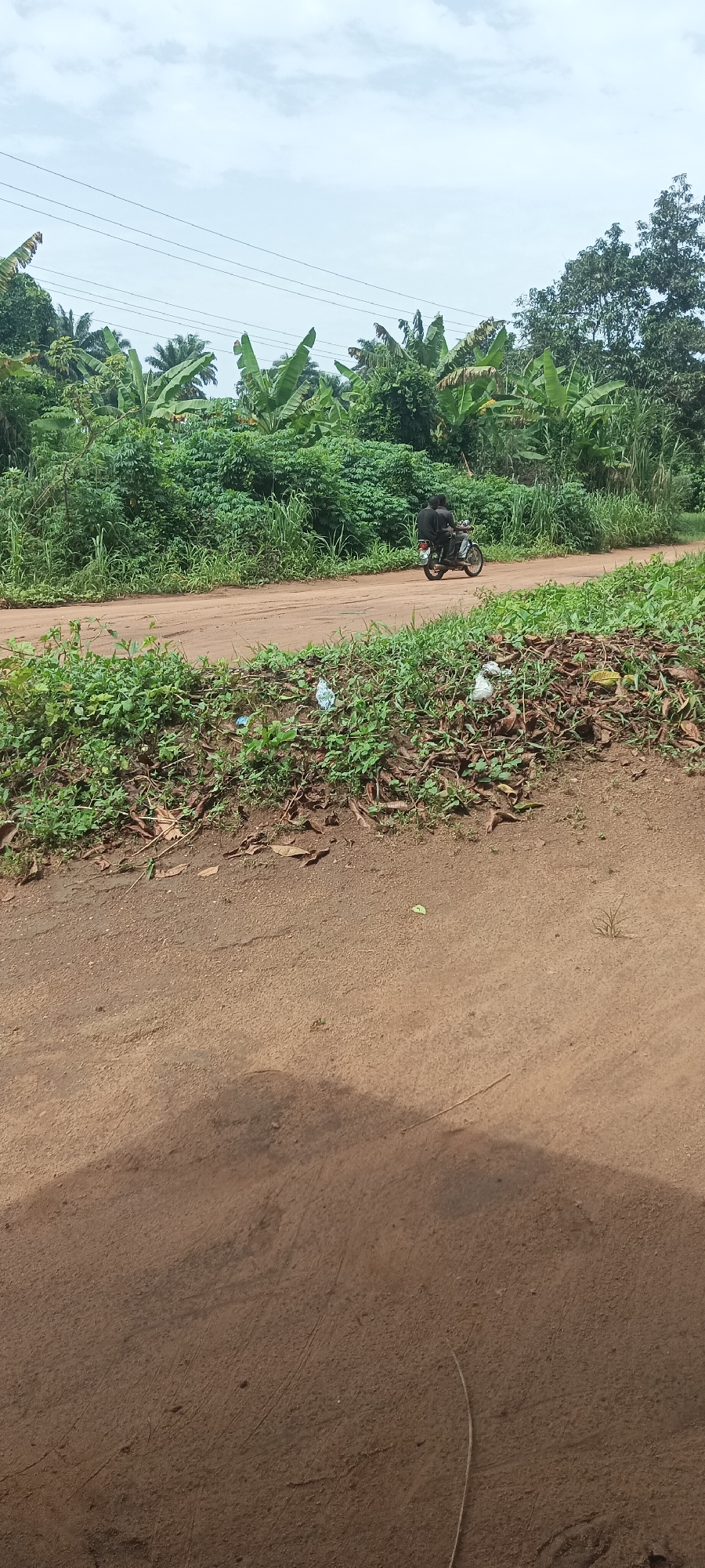 The image shows a dirt road in Amauzu, Nigeria. Dense green vegetation lines the road. Two people on a motorcycle are traveling along the road away from the viewer. There is trash and debris along the roadside. Power lines are visible in the background.