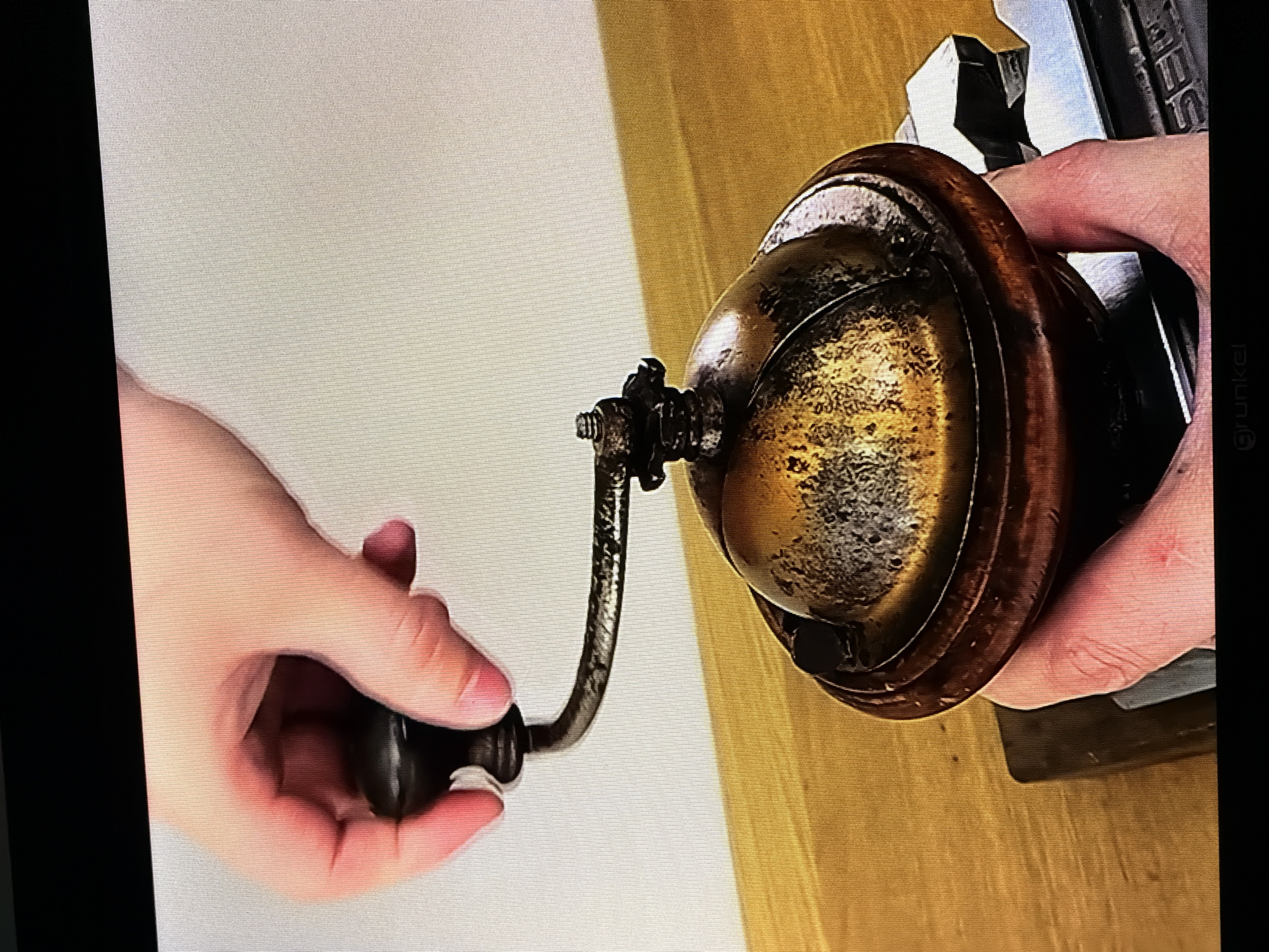A hand is shown operating a vintage coffee grinder. The grinder appears to be made of wood and metal, with a brass-colored top. The hand is holding the crank of the grinder and is in the process of turning it. The background is a plain white wall, and the grinder is placed on a wooden surface. The scene is brightly lit, suggesting it is daytime. There is no indication of Castillo Bajo-conejito, Spain in the image.