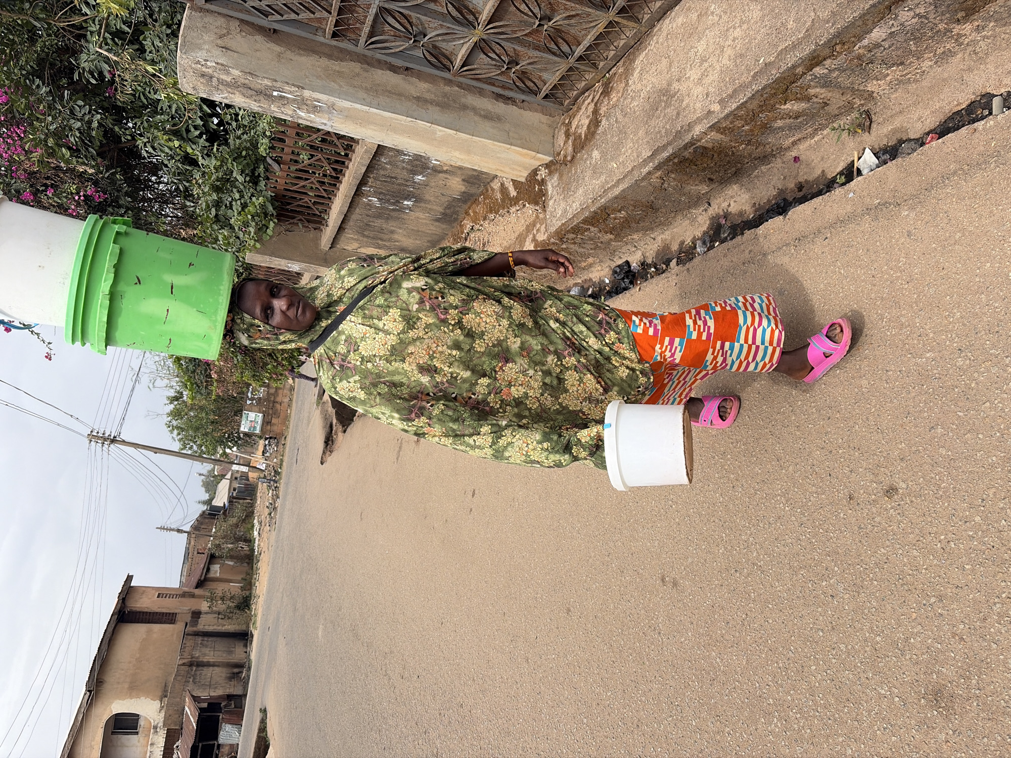 A woman is walking down a street in Jos, Nigeria, balancing two buckets, one white and one green, on her head. She wears a headscarf and a floral patterned green dress over an orange patterned skirt. She wears pink sandals. She has one bucket in her hand, and is on a gray road, next to a sidewalk with foliage on it. A building is on the left side of the frame. Power lines appear in the sky.