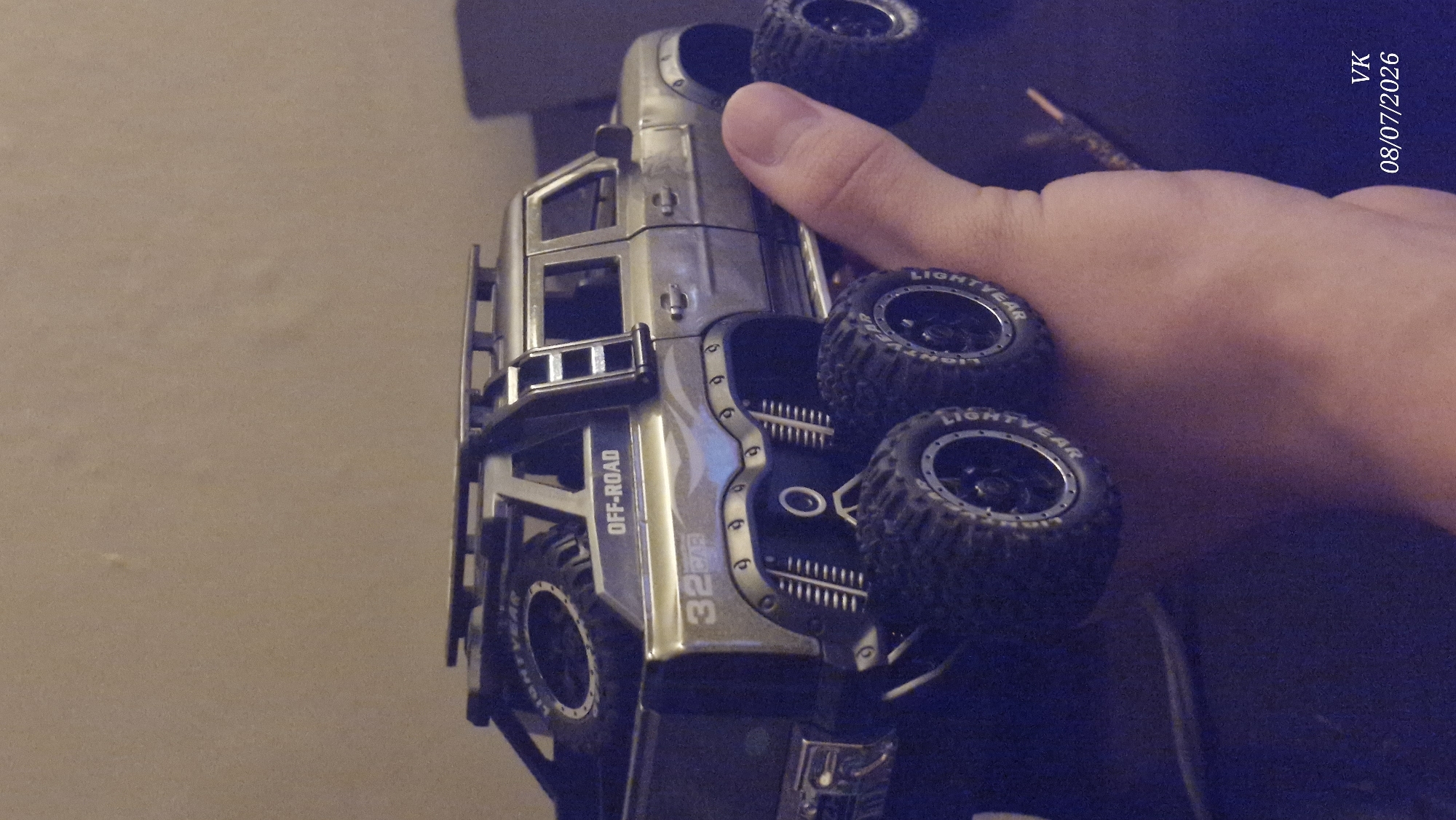 A toy off-road vehicle with six large wheels, a spare tire on the back, and a ladder on the side is held in a person's hand. The vehicle is silver with "OFF-ROAD" and "32 GTR" written on its body. The background is a plain, light-colored wall. A timestamp "VK 08/07/2026" is visible in the bottom right corner. There is no indication of Vrindavan, India in the image.