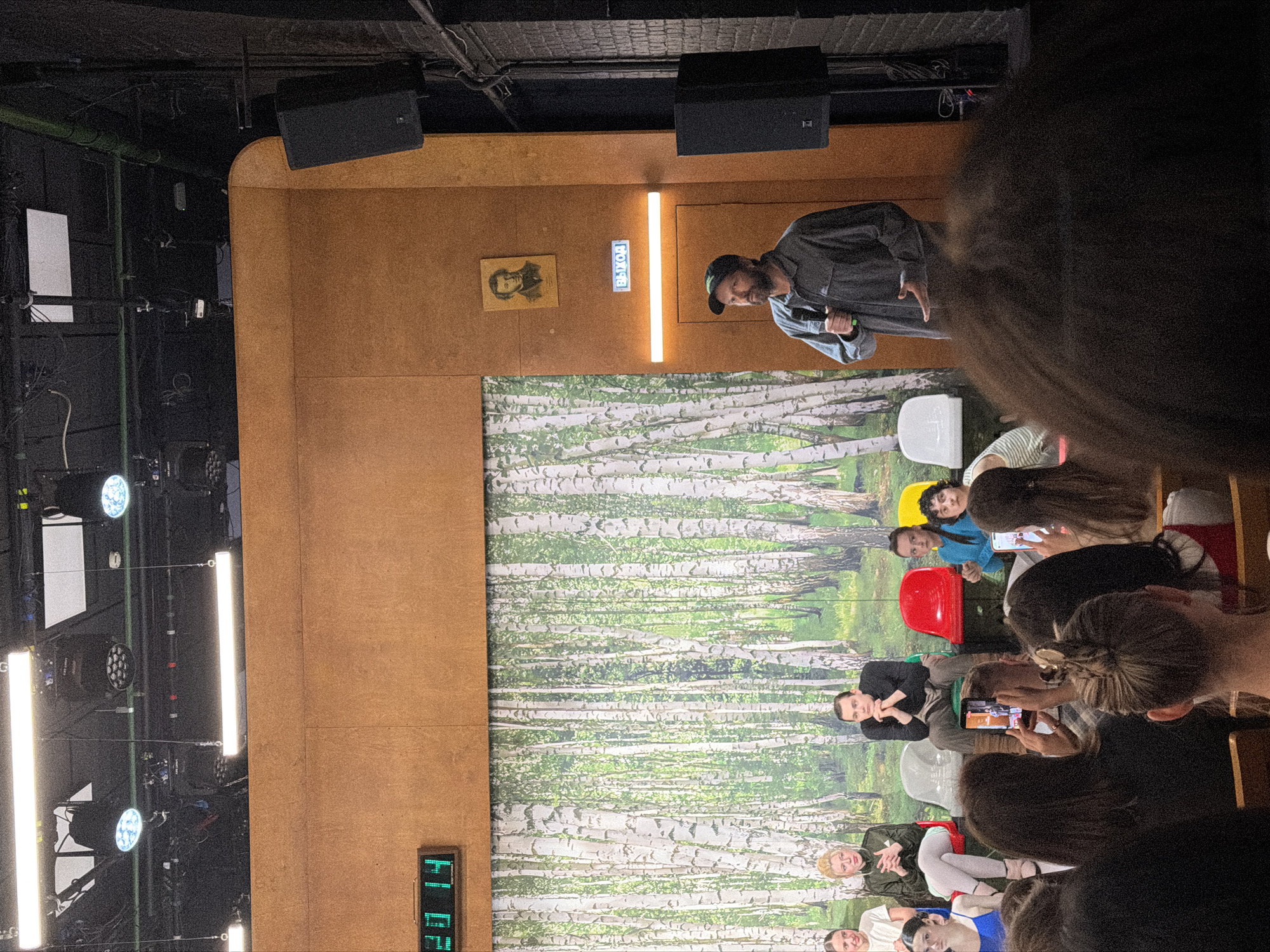 This image captures an indoor event, likely a presentation or performance, taking place in what appears to be a theater or lecture hall. The scene is dominated by a stage area on the left and an audience seated in front of it on the right.
On the stage, a man with a beard and wearing a cap and a dark jacket is speaking into a microphone. He stands in front of a large backdrop featuring a vibrant image of a birch forest. The stage itself is constructed with light brown wooden panels. Above him, a bright horizontal bar light illuminates the area. To his left, mounted on the wooden wall, is a framed portrait of an older man with a serious expression, and next to it, an illuminated sign reads "ВЫХОД" (VYKHOD), which is Russian for "EXIT," confirming the Moscow, Russia context. Above the stage are industrial-style ceilings with various stage lights and large black speakers.
To the left of the stage, several vertical bar lights and spotlights are visible, indicating professional lighting setup for the event. A digital clock on the bottom left of the wooden stage wall displays "20:14," suggesting the event is taking place in the evening.
The audience, positioned partially in shadow, consists of several individuals seated on various colored chairs (red, white, yellow, gray). Many audience members are looking towards the stage, and some are holding up mobile phones, seemingly recording or photographing the speaker. Their expressions range from attentive to casual observation. The hair and upper body of an audience member are prominently in the foreground, partially obscuring the view of others.