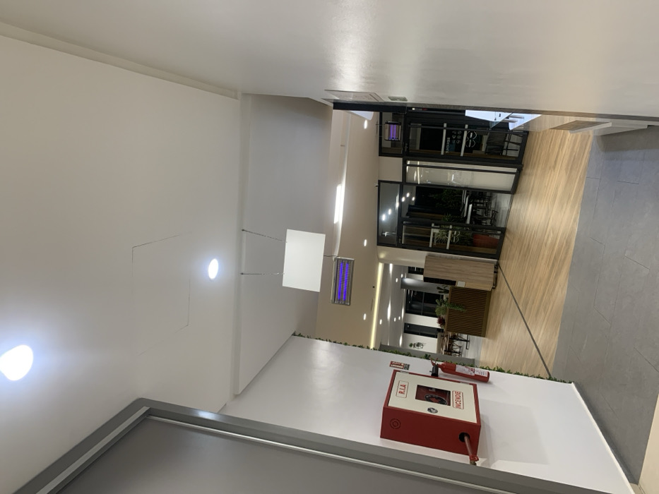This image depicts an interior hallway or common area located in Khemisset, Morocco. The scene is illuminated by overhead recessed LED lights and appears well-lit.
In the foreground, a dark grey tiled floor transitions into a lighter wood-patterned floor extending deeper into the space. A white wall on the left features a red fire hose cabinet labeled "R.I.A" and "INCENDIE" in white text, with a hose visibly connected. A red fire extinguisher is mounted immediately to the right of the cabinet. The upper left portion of the frame shows a grey door frame or architectural element.
The white ceiling displays a visible rectangular access panel and a white rectangular sign suspended by two thin wires. Further into the scene, a ceiling-mounted insect trap emitting a purple-blue light is visible.
To the right, another white wall with a partial view of a counter or display unit is present. The midground opens into a larger area with various architectural elements, including wooden slatted structures and light-colored wooden panels. Glass doors lead into adjacent rooms; one reveals tables and chairs, suggesting a dining or café area. Another glass-paneled entrance displays reversed text and graphics, possibly "BENCHE" and an optician's logo. A second blue-light insect trap hangs near this entrance in the background. No individuals, specific actions, or direct interactions are visible within the frame.