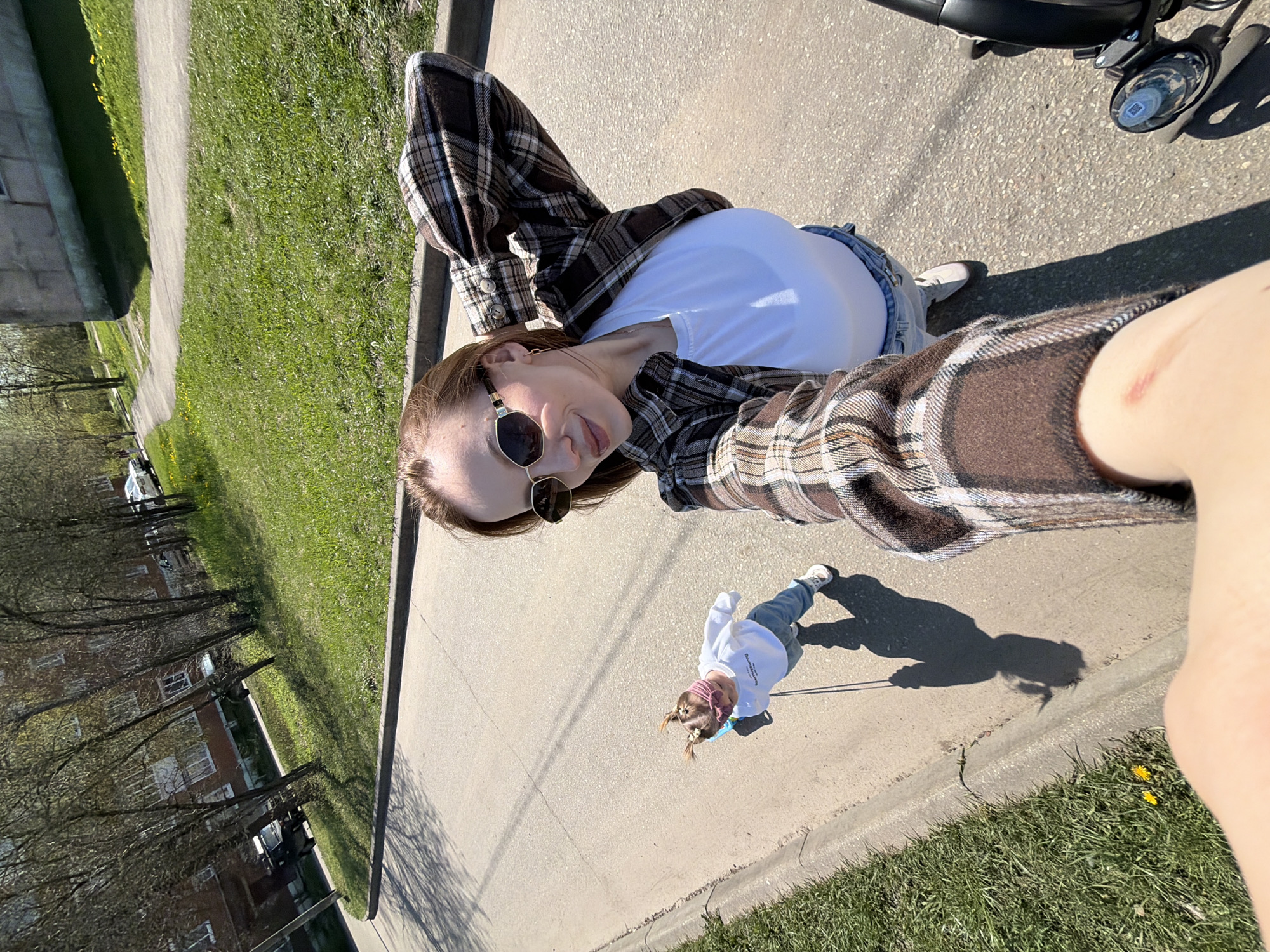 A woman in a white T-shirt and plaid shirt is taking a selfie outside in the city of Reshetnikovo, Russia. She is on a paved surface, possibly a sidewalk or road. A young child, wearing jeans and a white shirt with a pink hair band, is walking on a leash in front of her. A motorized vehicle is partially visible at the top of the frame. To the left, there are trees and buildings, likely residential, adjacent to a grassy area.