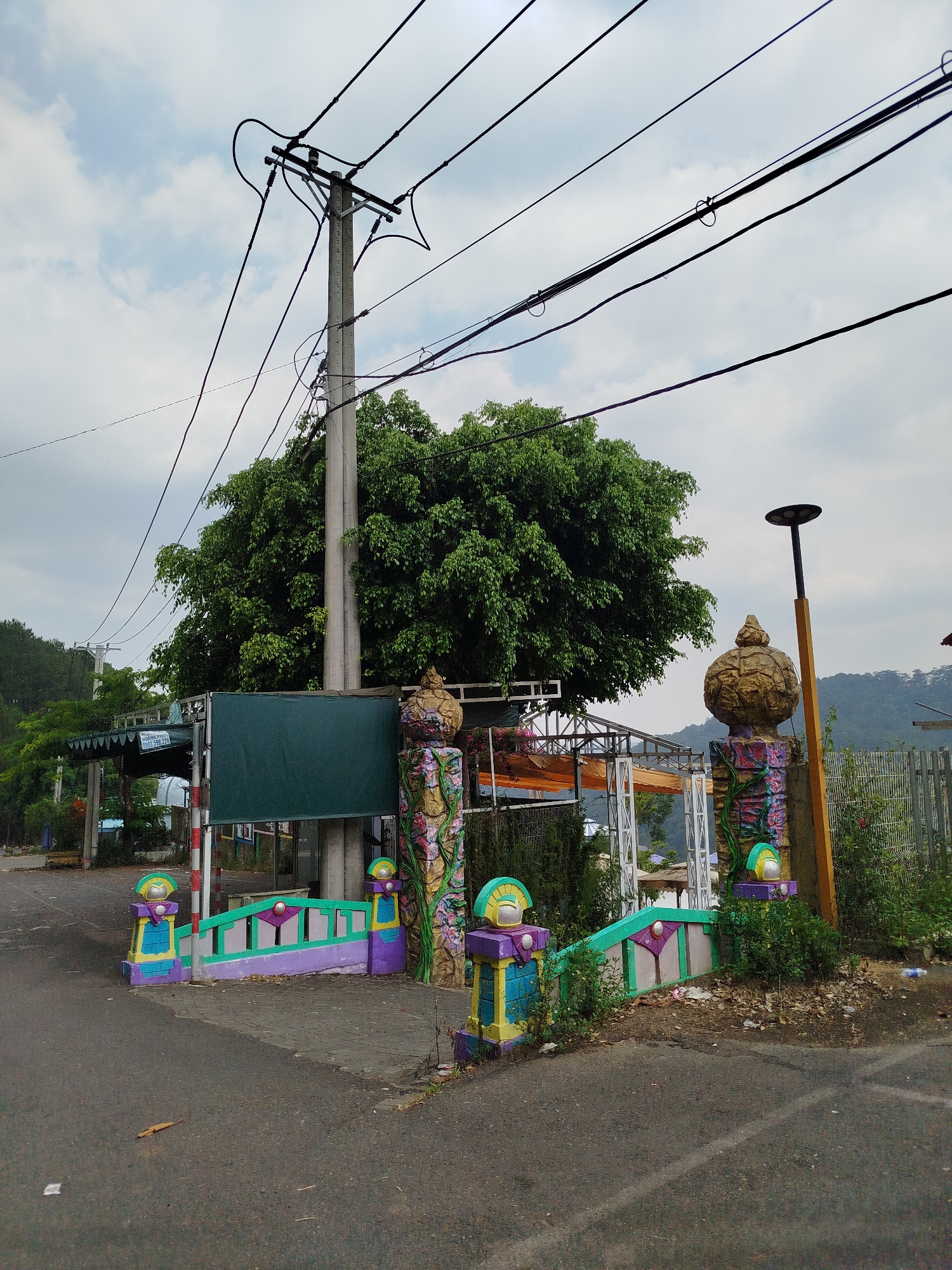 The image shows a paved road leading to a colorful, ornate gate structure flanked by decorative pillars. Behind the gate is a sheltered area and trees. An electrical pole with numerous wires is prominent in the background. The scene is likely located in the city of Da Lat, Vietnam, as the gate exhibits styles and colors prevalent in the region. No people are visible.