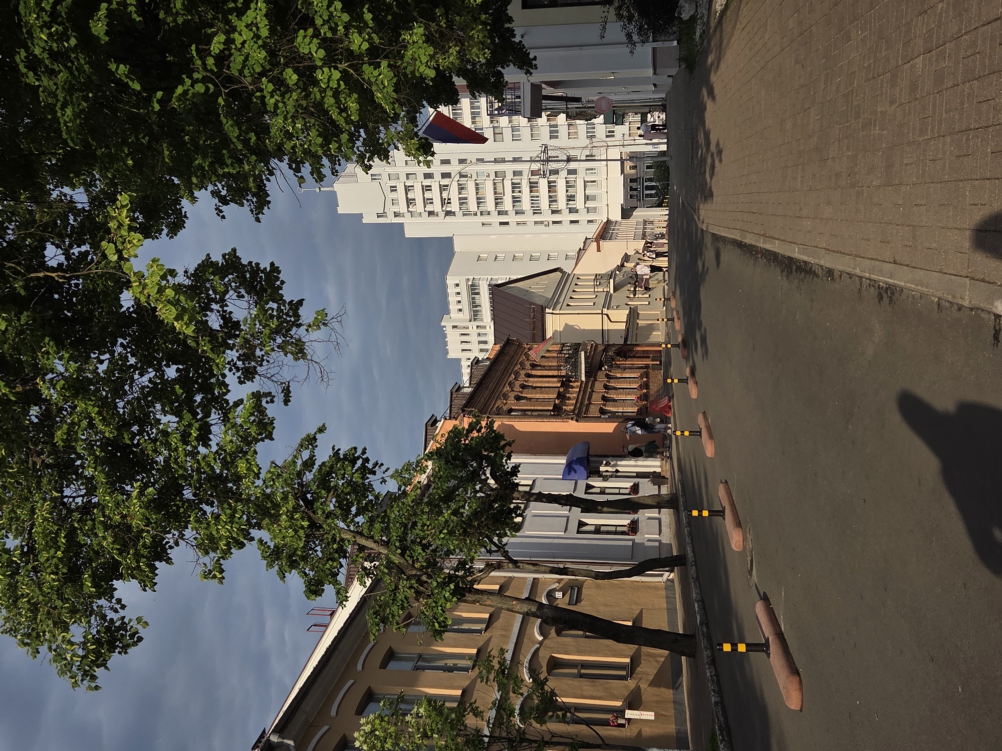 Here is a detailed description of the image:
The image depicts a street scene in an urban environment. The view is oriented along a street lined with buildings on both sides, leading into the distance. On the left side of the street, various buildings of different colors and architectural styles are visible. In the background, a tall white building with numerous windows rises above the other structures. Green trees with dense foliage are positioned along the street, partially obscuring the buildings and casting shadows on the scene.
The street itself is paved with asphalt and has a row of bollards placed along the edge, presumably to separate the street from the sidewalk or pedestrian area. On the right side of the street, a paved walkway is visible, slightly elevated from the road surface. Several people can be seen walking or sitting along this walkway, adding a sense of activity to the scene.
The sky is overcast, with clouds creating a soft, diffused light that affects the overall color and contrast of the image. The orientation of the photograph is rotated at an angle, making the street appear to run diagonally across the frame. The location of the city is unknown.