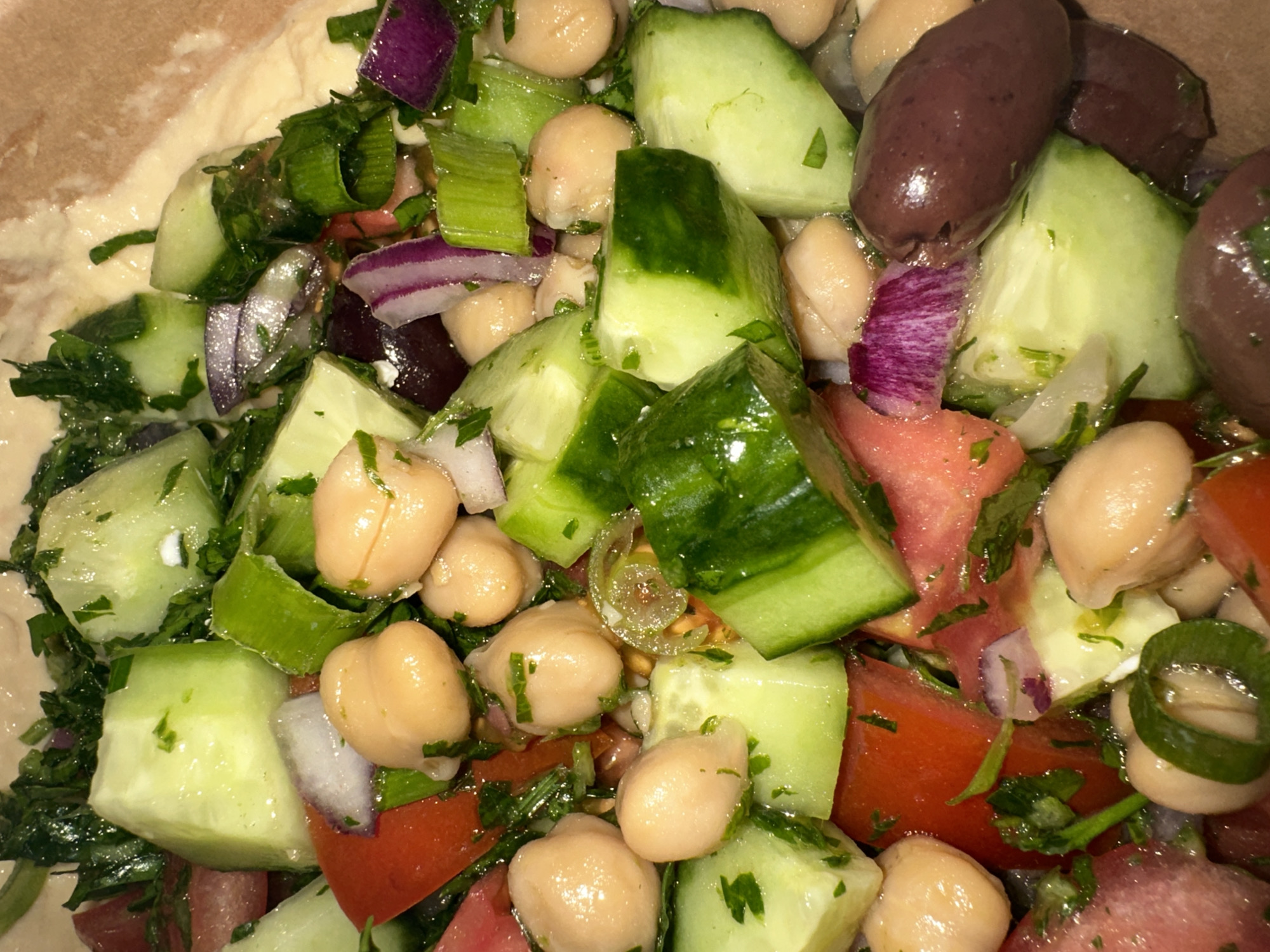 The image presents a close-up view of a salad. The salad contains chopped cucumbers, tomatoes, red onion, scallions, olives, and chickpeas, all mixed with parsley. The salad is coated in a glossy liquid, which may be a dressing or oil. The presence of hummus is visible at the upper-left corner of the image. The salad appears to be in a bowl or container. There is no information to associate this image with the city of Sofia, Bulgaria.