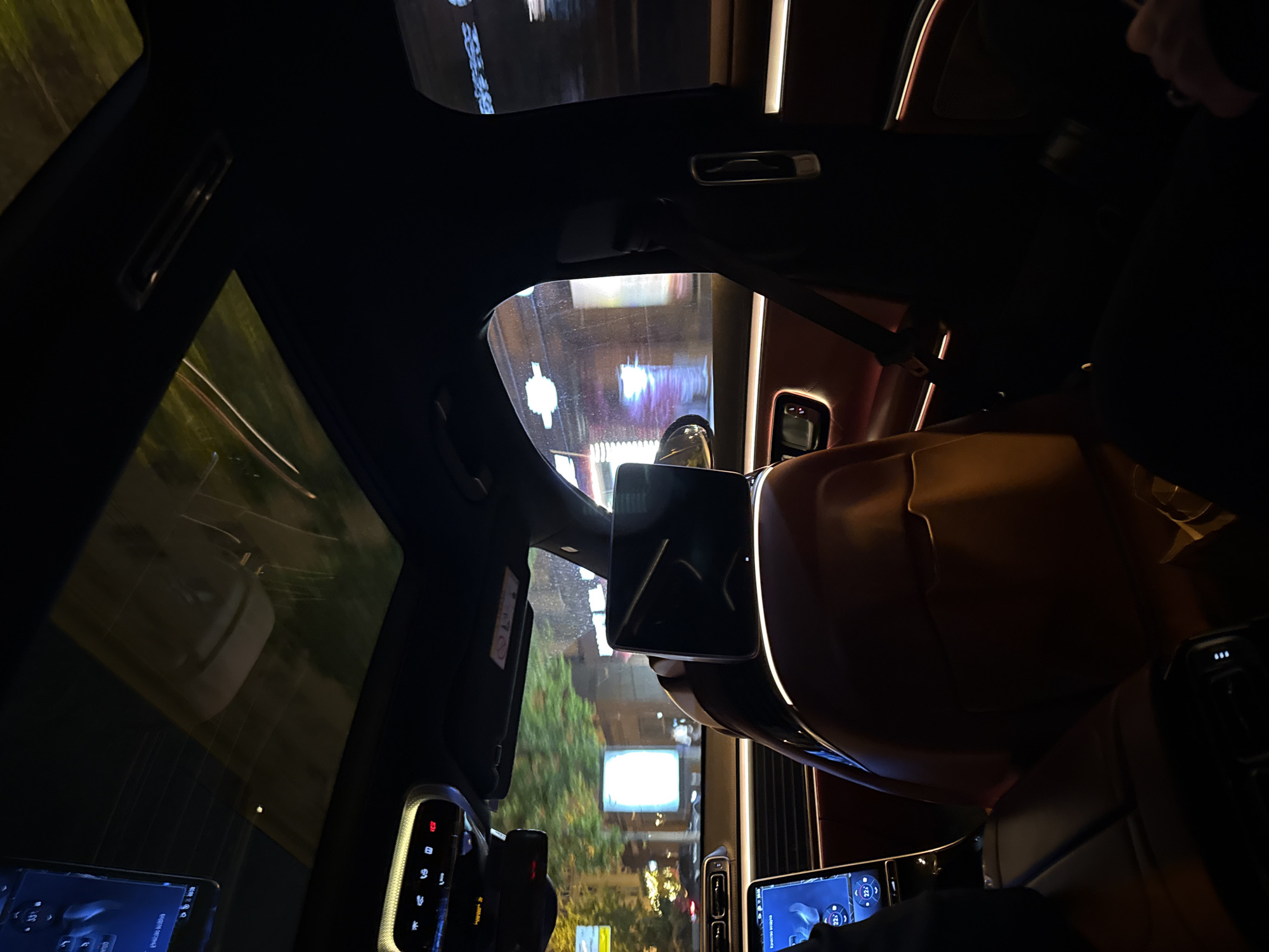 From the vantage point within a vehicle at night, a rainy street scene in Moscow, Russia, is captured. The vehicle's interior, featuring displays and controls, is dimly lit. Reflections of urban lights are visible on the wet pavement outside, including a prominent rectangular billboard.