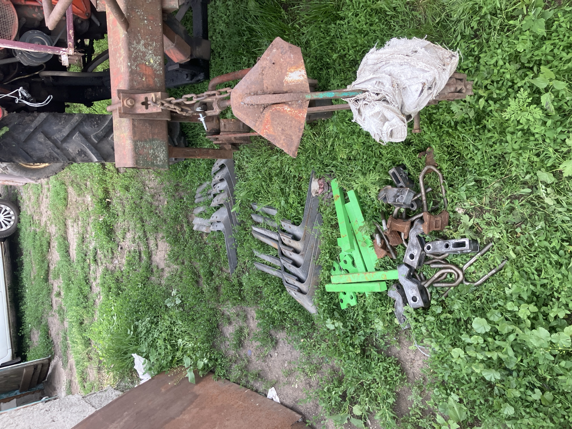 The image shows a collection of agricultural equipment and parts scattered on the grass in a yard, likely in Vovkivka, Ukraine. A tractor's rear tire and part of its frame are visible on the right side of the image, suggesting the equipment may be associated with it. Several plowshares, made of metal and some painted green, are laid out on the ground. Other metal components, including U-bolts and various brackets, are also visible, some appearing rusty. A rusty, triangular metal piece, possibly an anchor or part of a plow, is suspended from a chain attached to the tractor. A white, torn burlap sack is wrapped around the lower part of this suspended metal piece. A portion of a white vehicle and a rough-hewn wooden structure are visible on the left side, indicating a rural or agricultural setting. The ground is covered with green grass and some dry soil.