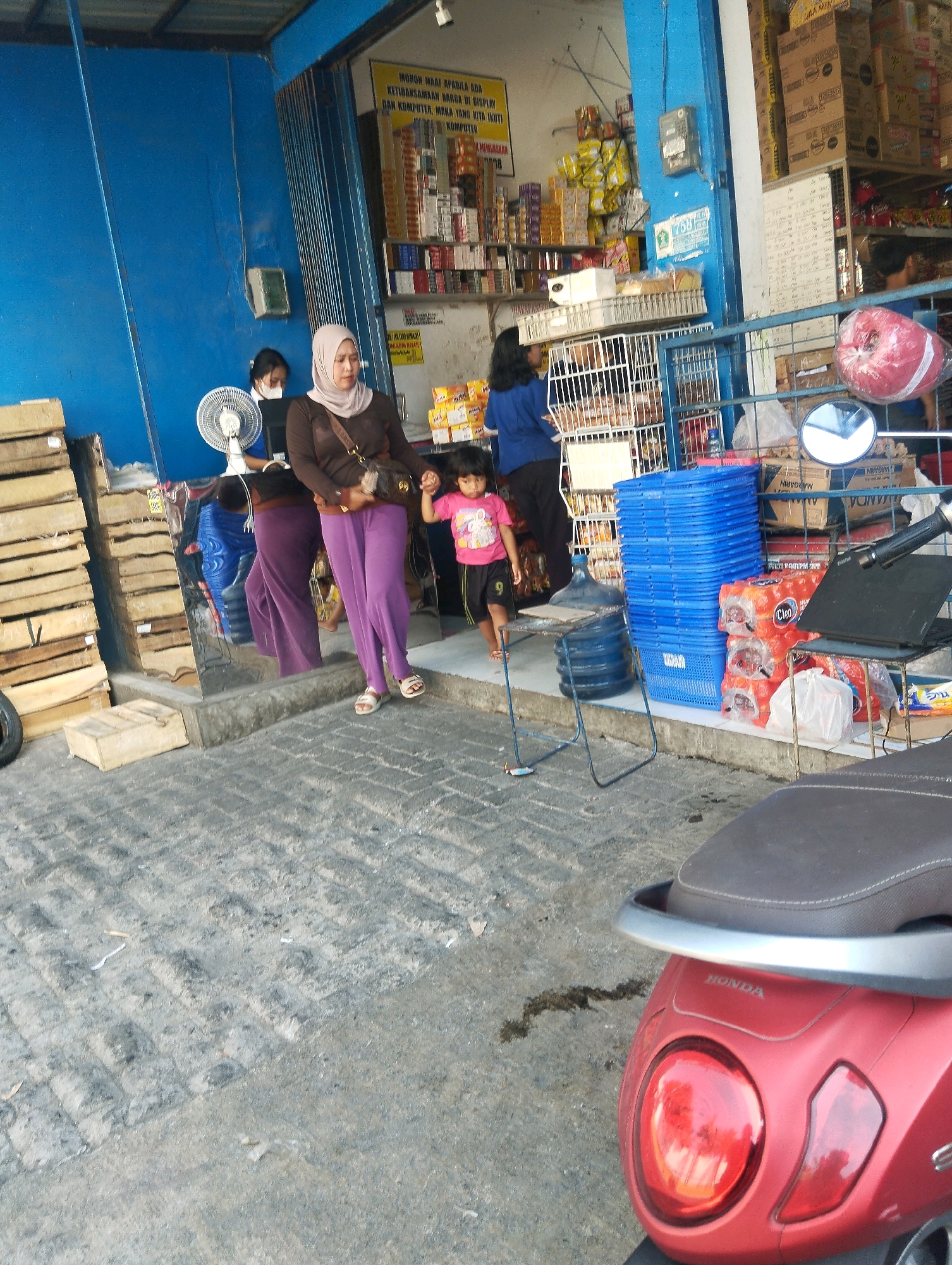 The image captures a street scene in front of a shop in Malang, Indonesia. A woman wearing a light-colored hijab and a brown top, along with dark purple pants, is holding the hand of a young child wearing a pink t-shirt and black shorts. They are walking on a paved area in front of a blue-walled establishment that appears to be a retail store. Inside the store, shelves are stocked with various goods, including boxes and packaged items. A white fan is visible near the entrance, and several blue plastic baskets are stacked outside. A large blue water jug is also present. The foreground shows a red scooter, partially visible, with the "HONDA" logo on its rear. The ground is a textured gray pavement. The overall scene suggests a typical commercial area in the city.