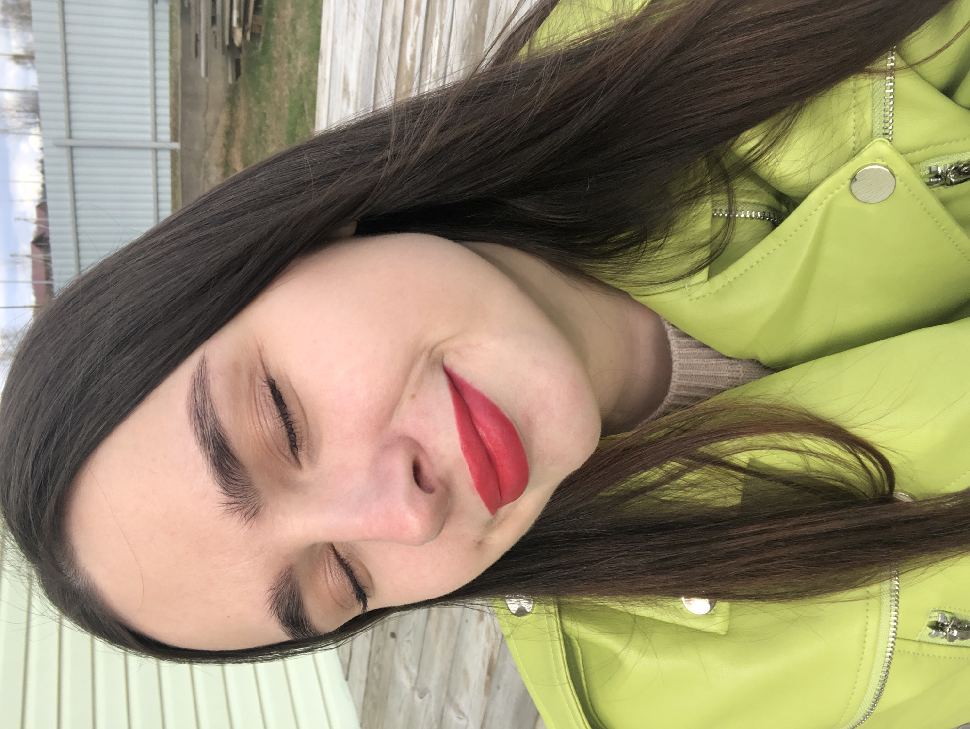 A young woman with dark brown hair is captured in a close-up, slightly angled portrait. Her eyes are closed, and she is wearing bright red lipstick. She is wearing a lime green jacket with silver zipper and button details. The background shows a wooden fence and glimpses of greenery. The location is possibly in Noviy, Russia.