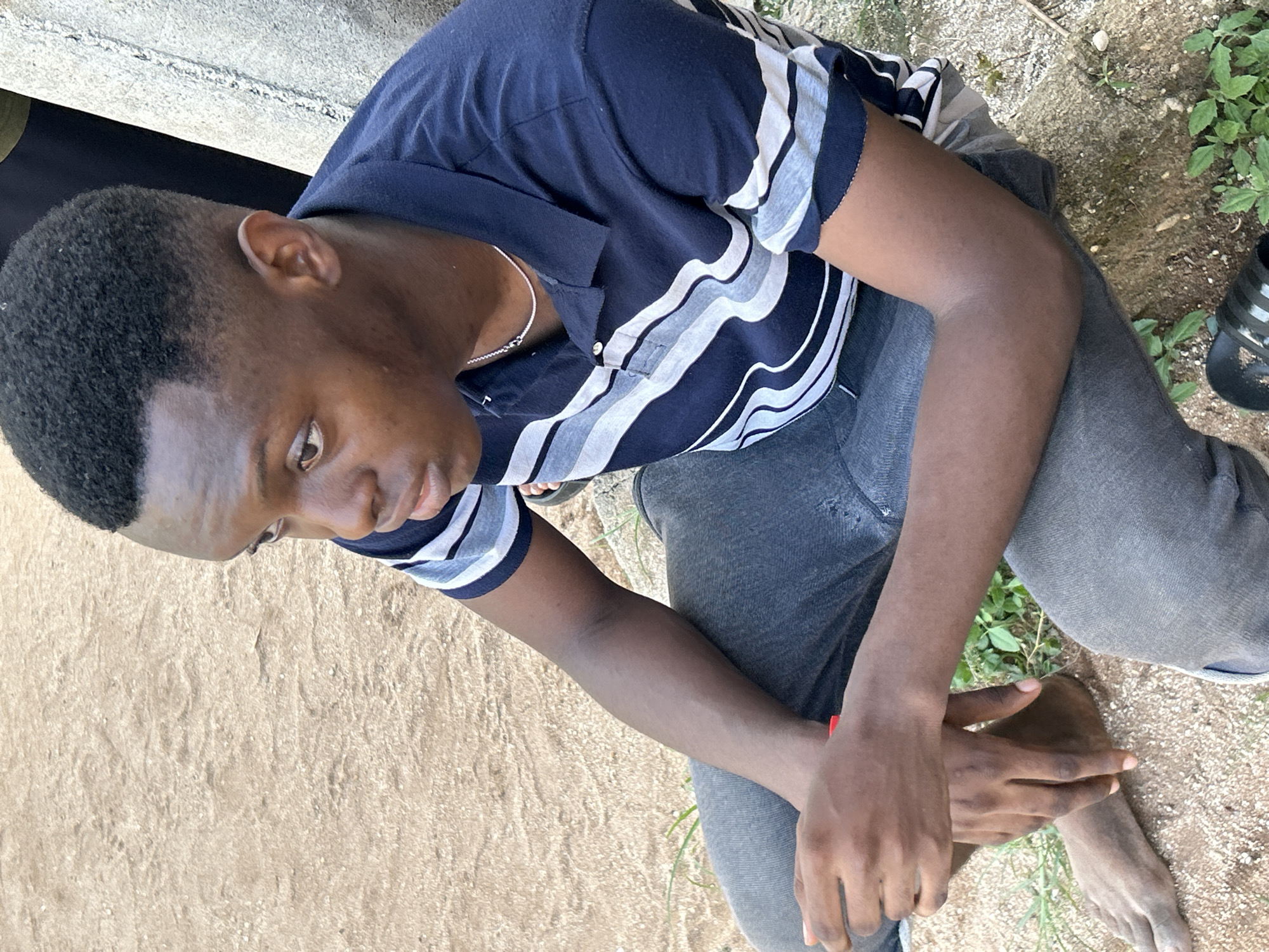 The image shows a young man sitting on a concrete surface in Ihiagwa, Nigeria. He is wearing a navy blue polo shirt with white and gray horizontal stripes, and gray jeans. He is looking down and his hands are clasped together in front of him. He is also wearing a silver chain. There is a concrete structure behind him, and some greenery and footwear are visible nearby. The ground surrounding him is sandy.