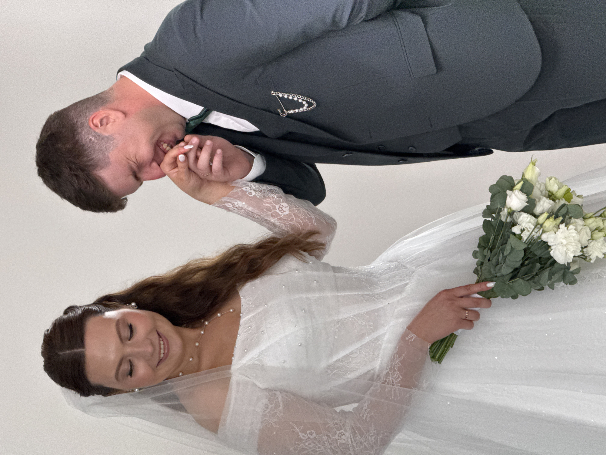 An indoor photograph captures a joyful wedding moment between a bride and groom against a plain, light background. The image is oriented vertically, showing the groom positioned higher on the left, leaning down towards the bride who is positioned lower on the right.
The groom, with short dark hair, wears a dark green or grey suit jacket over a white shirt and a dark green tie. A decorative silver lapel pin, shaped like a 'D' or a stylized safety pin adorned with small pearls or crystals, is visible on his left lapel. He is smiling broadly, with his mouth open, as if playfully biting or kissing the bride's hand, which he holds gently. His expression conveys clear affection and happiness.
The bride, with long brown hair styled in a half-updo, also smiles widely with closed eyes, indicating a moment of pure bliss. She wears a white wedding gown featuring long, sheer lace sleeves with intricate patterns. The dress fabric also appears to have subtle sparkles or scattered pearls. She accessorizes with a delicate pearl necklace and matching pearl drop earrings. A long, sheer white veil frames her face and flows down her back.
In her right hand, the bride holds a wedding bouquet composed of white flowers, likely eustoma or lisianthus, mixed with green eucalyptus leaves. A simple gold wedding ring is visible on the fourth finger of her left hand, which the groom is holding. Her fingernails are painted white.
The lighting is bright and even, suggesting a controlled studio environment or a well-lit indoor space. The focus is entirely on the couple and their interaction, highlighting their emotions of happiness and intimacy during what appears to be a wedding photoshoot. There is no visible text in the image. The setting does not provide specific cues for Chaikovsky, Russia, beyond the general context.