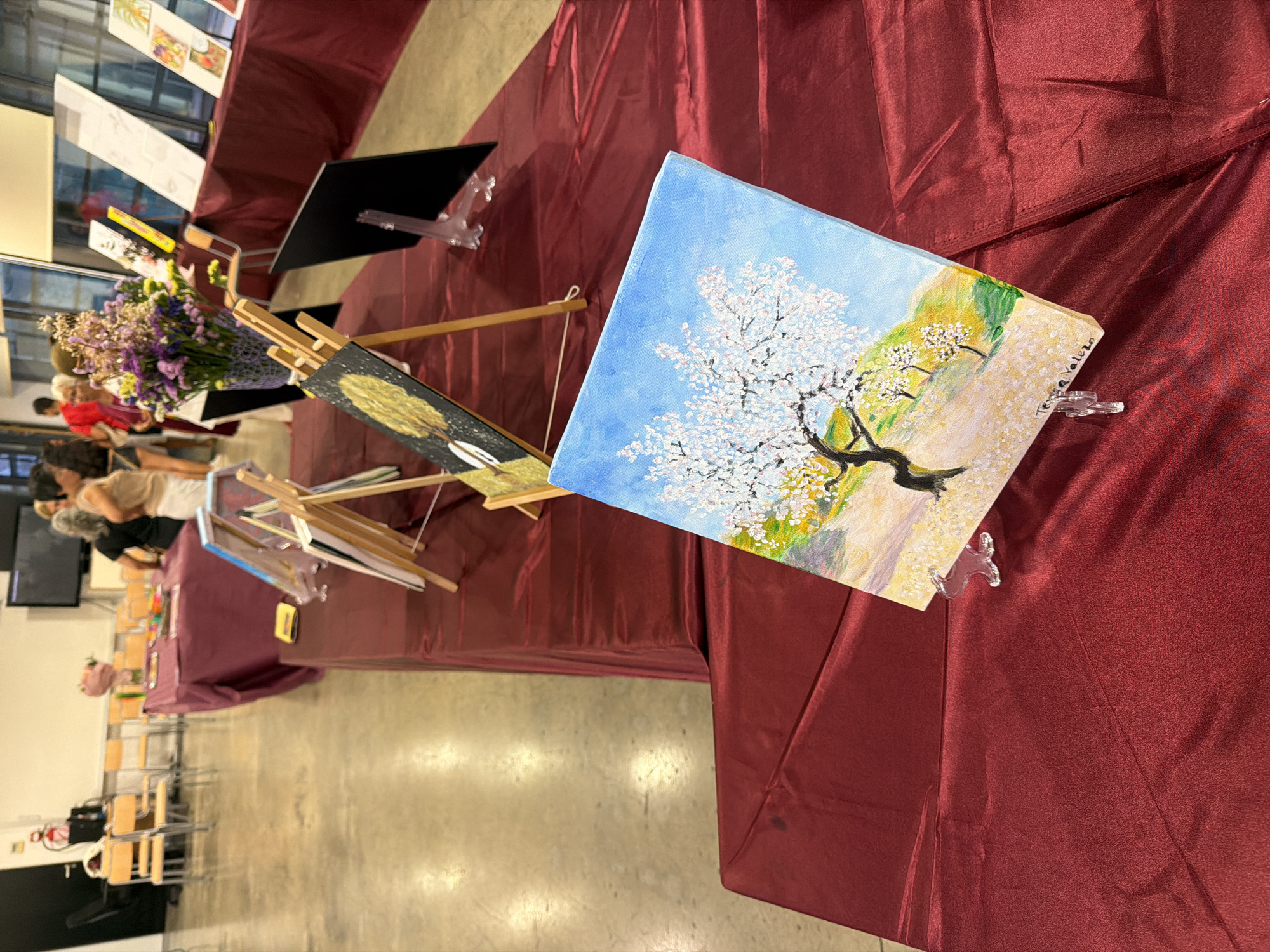 In Elche, Spain, this image captures a scene from an art exhibition. The focus is on a table draped with a burgundy cloth, displaying several pieces of art. In the foreground, a small painting of a flowering tree with a blue sky is propped up on a clear stand. Further back, more artworks are displayed on easels, including a painting of a tree with golden leaves against a dark background. A vase filled with colorful dried flowers sits on a distant table. In the background, chairs are arranged around tables covered in burgundy cloths, and several people are visible, appearing to browse the artwork. The environment seems to be an indoor exhibition space with concrete flooring.