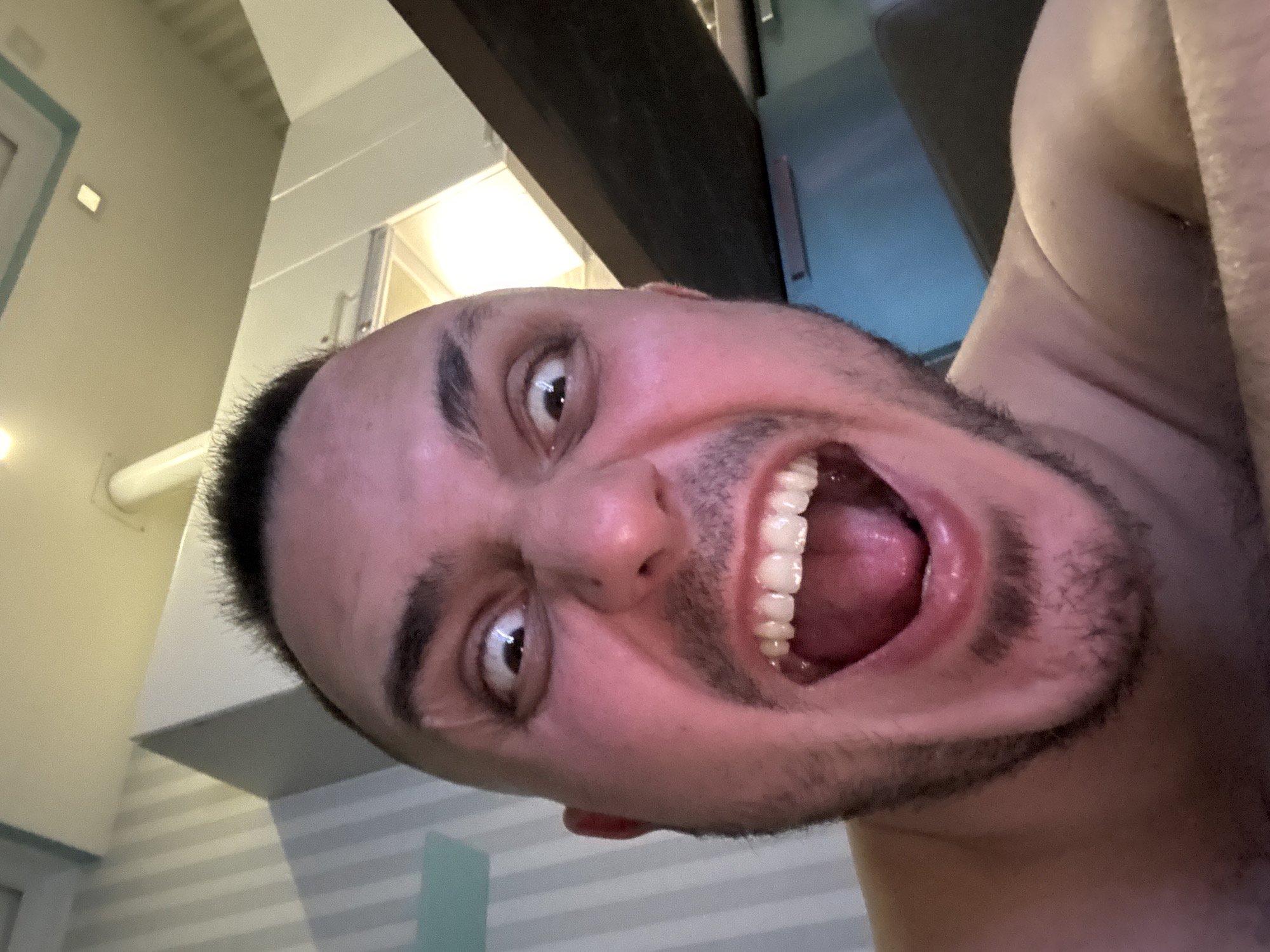 A close-up selfie of a man with dark hair and a beard. His mouth is open wide, revealing his teeth and tongue. He appears to be indoors, with a white striped wall visible in the background. A light fixture is visible in the upper portion of the image.
The image has no discernible information about Saint Petersburg, Russia.