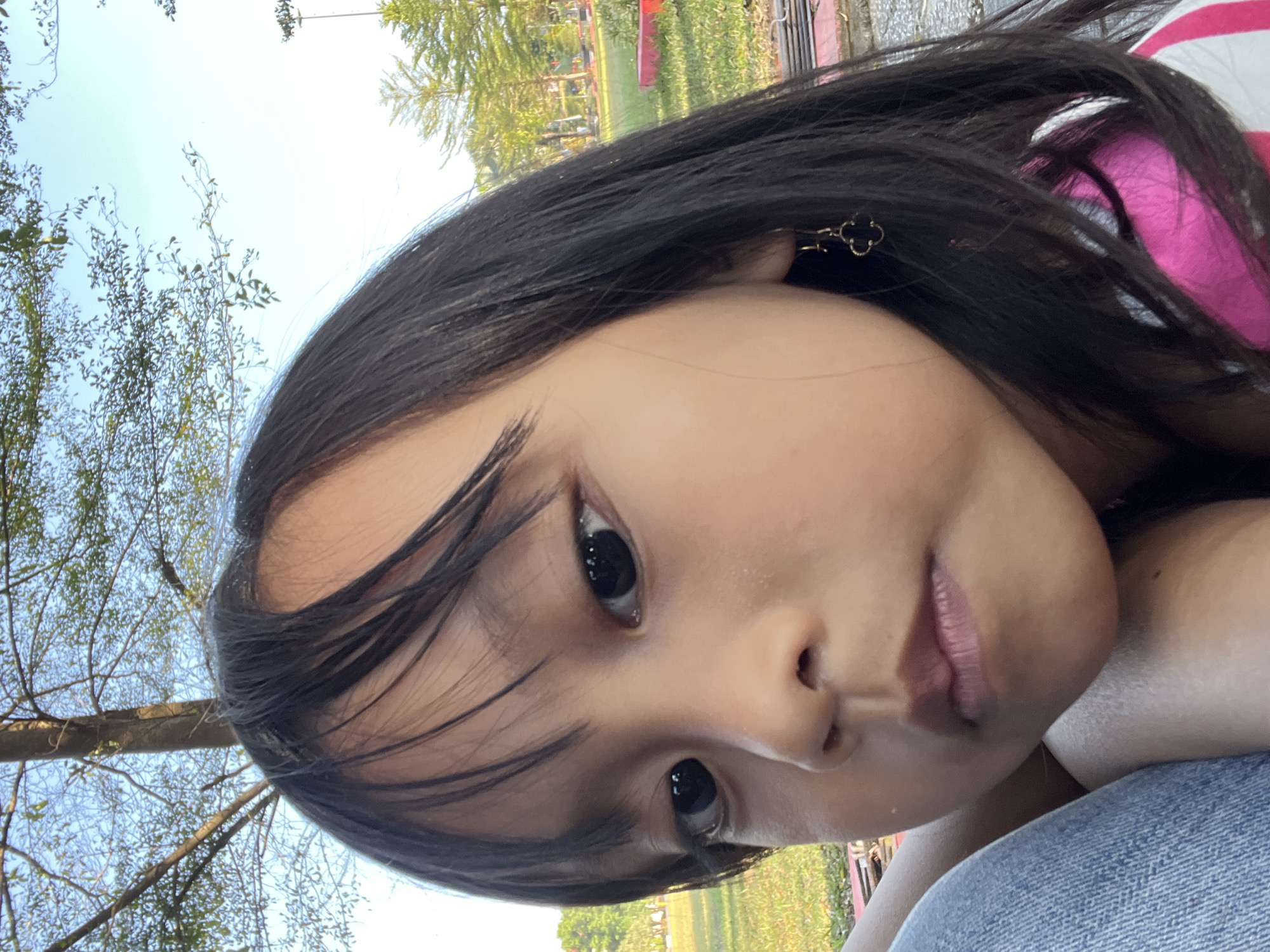 A close-up, low-angle selfie of a young East Asian girl is taken outdoors. She has long, dark hair, bangs, and is wearing a gold clover-shaped earring on her right ear. She is wearing a pink and white striped shirt and blue jeans. The background shows a park with trees and a bright blue sky. In the distance, there are more trees and a lamppost. The lighting suggests it is daytime. The girl is looking directly at the camera with a neutral expression. The scene is likely in the city of Purwodadi, Indonesia, given the park-like setting and vegetation.