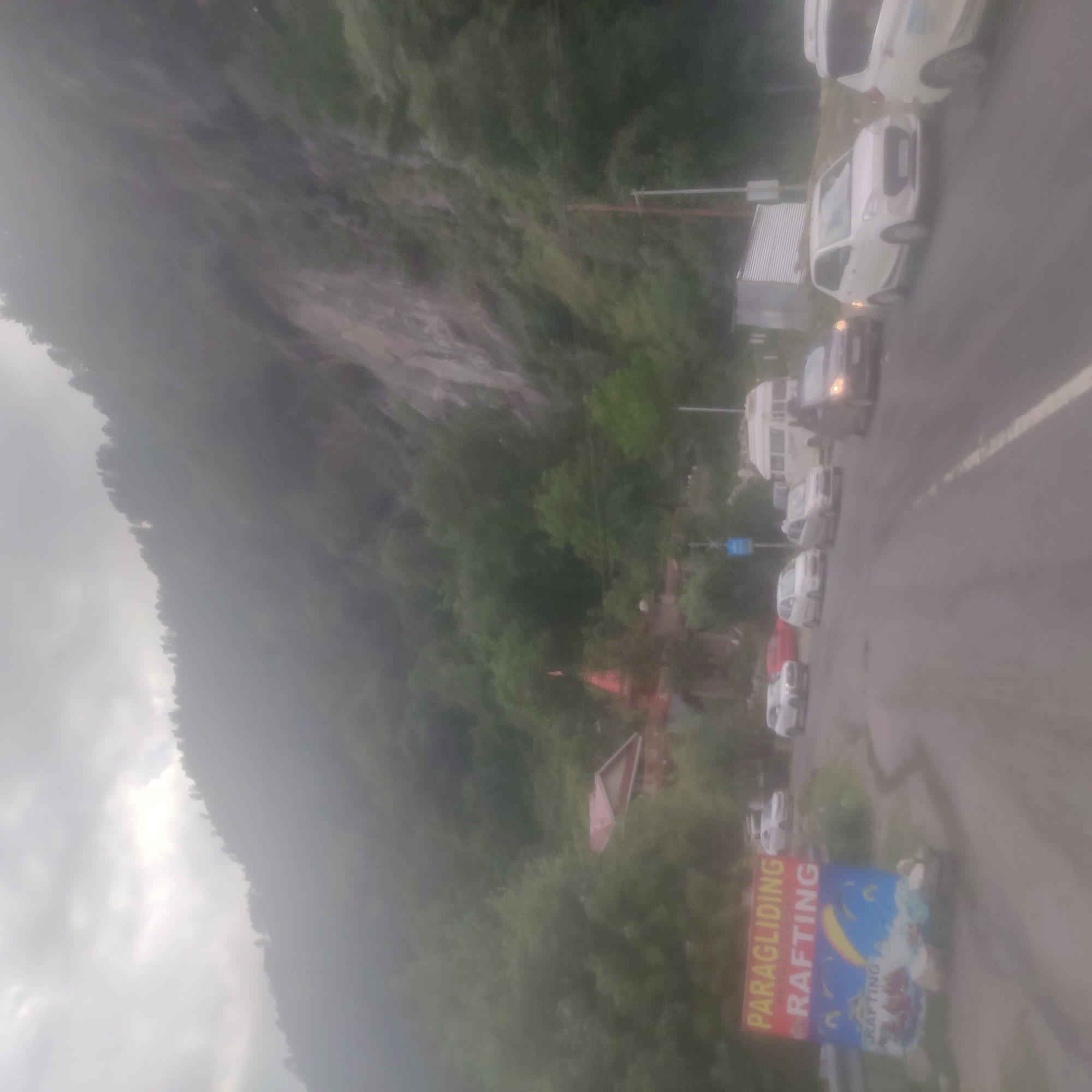 The image depicts a cloudy day scene on a road in or near Kalath, India, with a mountain backdrop. A sign advertising "PARAGLIDING RAFTING" is visible on the left. Several cars, including a white hatchback and a white van, are on the wet asphalt road, some in motion and some stopped. A red-roofed building, possibly a temple, is nestled among trees on the mountainside. The overall atmosphere is hazy, suggesting misty or rainy conditions.